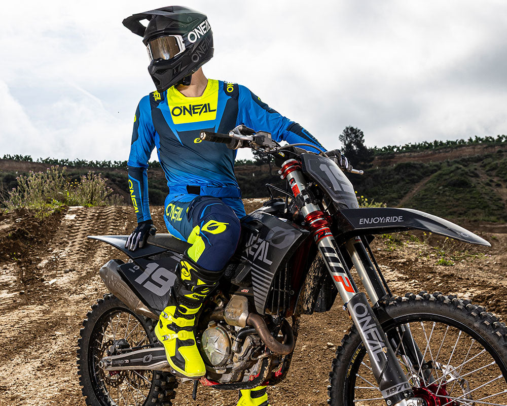 Ein Motocross-Fahrer in blauer und gelber O'Neal-Kleidung und mit Helm sitzt auf einem schwarzen Dirt Bike und blickt zur Seite auf eine unbefestigte Strecke mit Hügeln und Grün im Hintergrund.