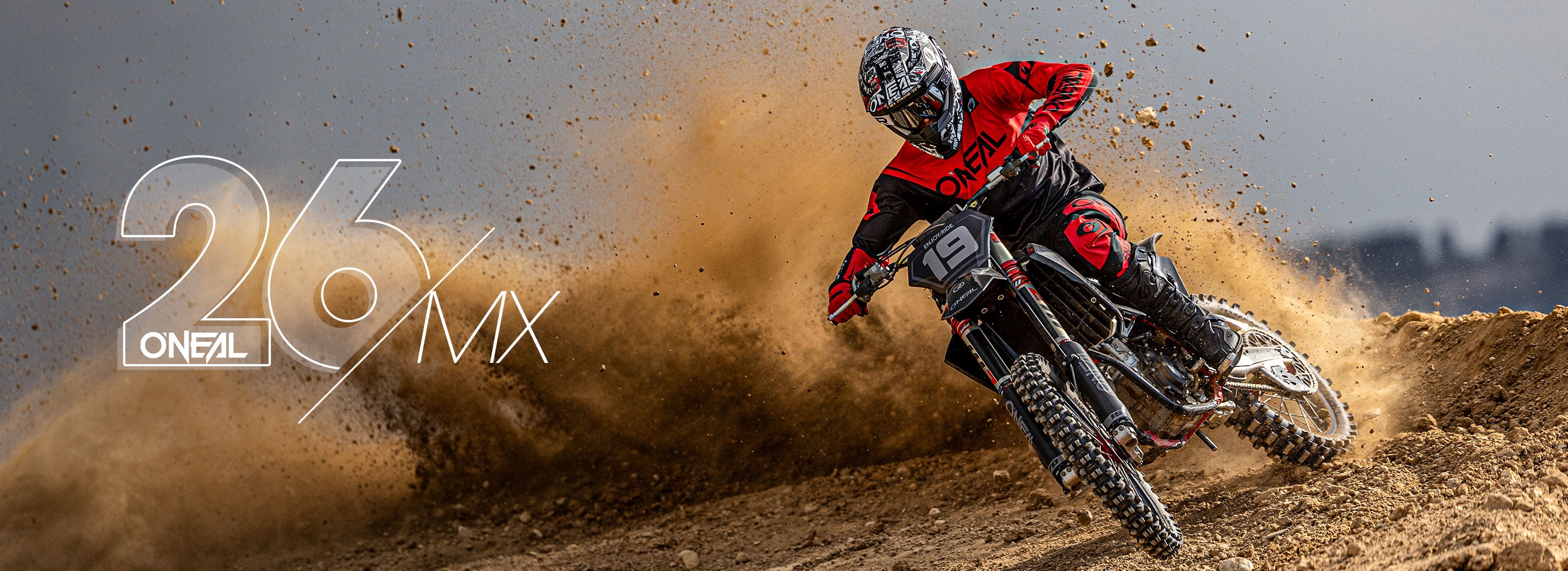 Ein Motocross-Fahrer in roter und schwarzer Kleidung fährt mit einem Dirt Bike um eine Kurve und wirbelt dabei Staub auf. Die Nummer 26 und das "O'NEAL MX"-Logo erscheinen auf der linken Seite des Bildes.