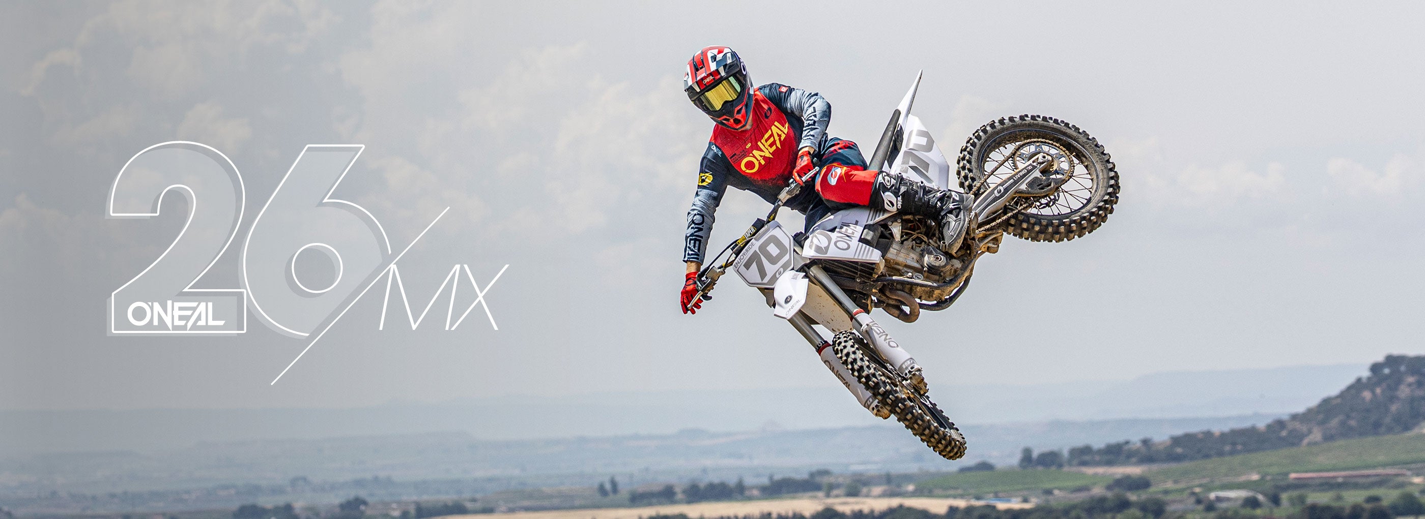 Ein Motocross-Fahrer in voller Montur vollführt einen Trick in der Luft auf einem Dirt Bike vor einer malerischen Kulisse im Freien, wobei der Text "26 O'NEAL MX" auf der linken Seite des Bildes eingeblendet ist.