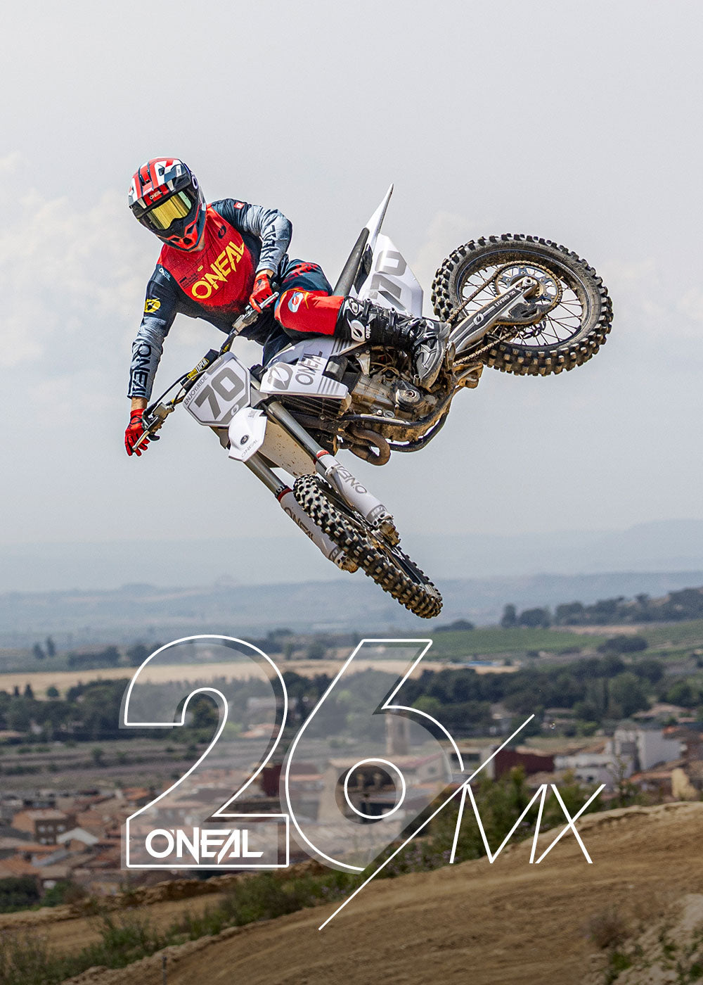 Ein Motocross-Fahrer in roter und blauer O'NEAL-Kleidung springt auf einem Dirt Bike durch die Luft, während im Hintergrund Felder, Gebäude und Hügel zu sehen sind. Im Vordergrund steht in großer weißer Schrift "26 O'NEAL MX".