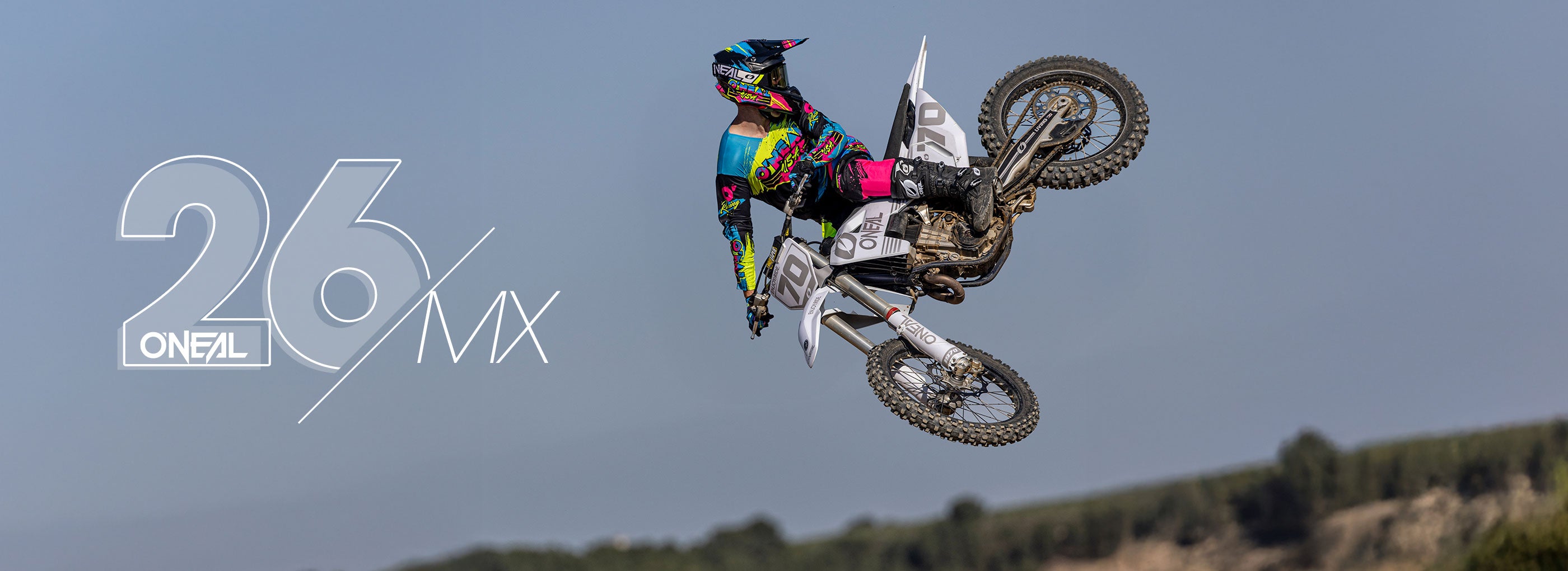 Ein Motocross-Fahrer in farbenfroher Ausrüstung vollführt einen Sprung in der Luft auf einem Dirt Bike vor einem klaren Himmel, wobei der Text "26 ONEAL MX" auf der linken Seite des Bildes angezeigt wird.