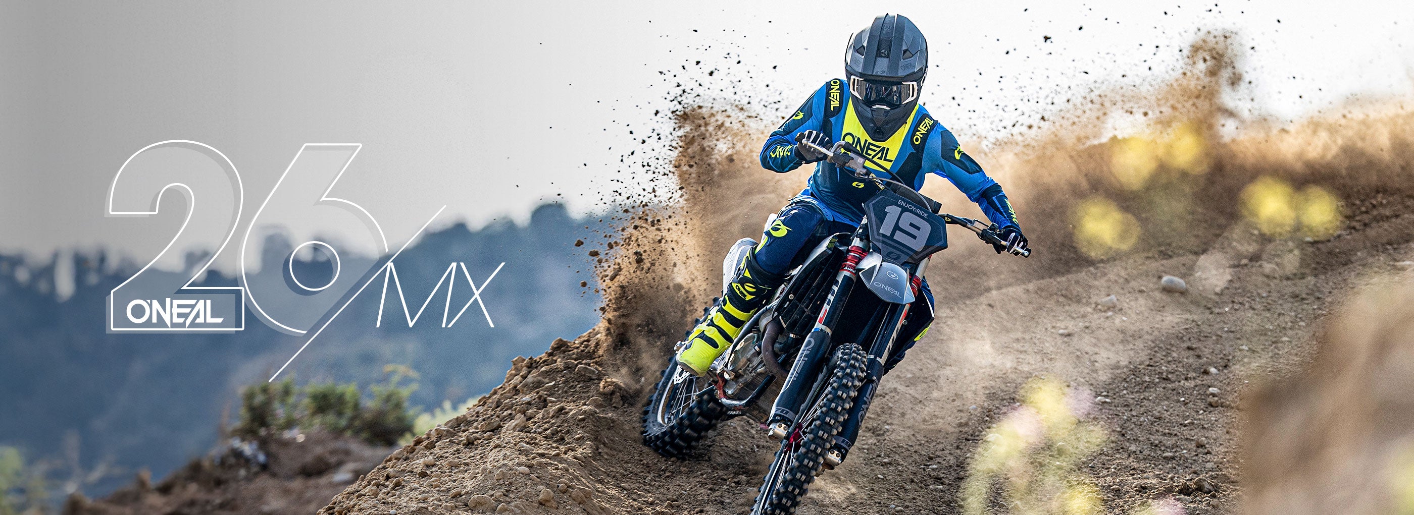 Ein Motocross-Fahrer in blauer und gelber Kleidung, mit Helm und Schutzbrille, rast auf einem Motorrad um eine Dirt-Track-Kurve und wirbelt dabei Staub auf. Der Text auf dem Bild lautet "2026 O'Neal MX.