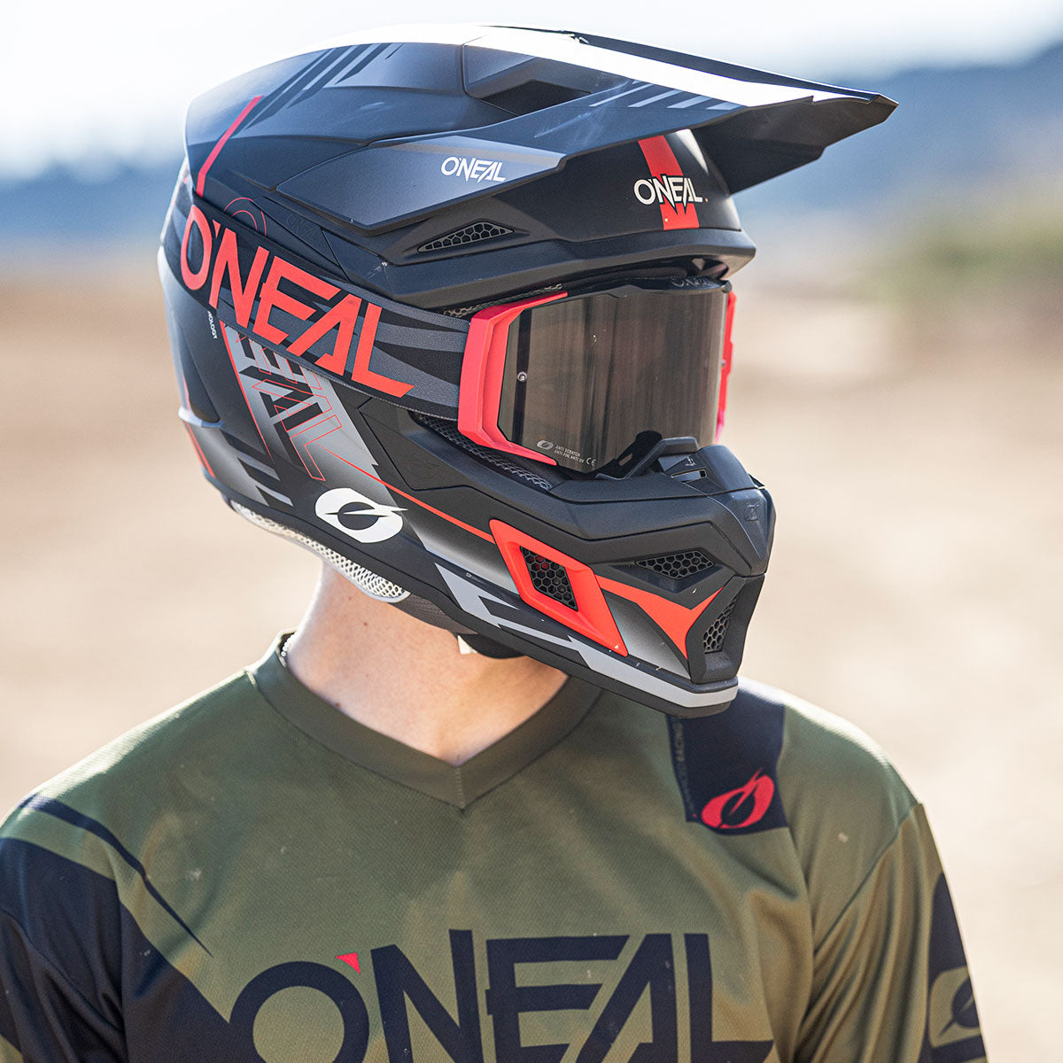Eine Person, die einen schwarz-roten O'Neal-Motocross-Helm mit Schutzbrille und ein passendes grün-schwarzes O'Neal-Trikot trägt, steht im Freien vor einem unscharfen Hintergrund.