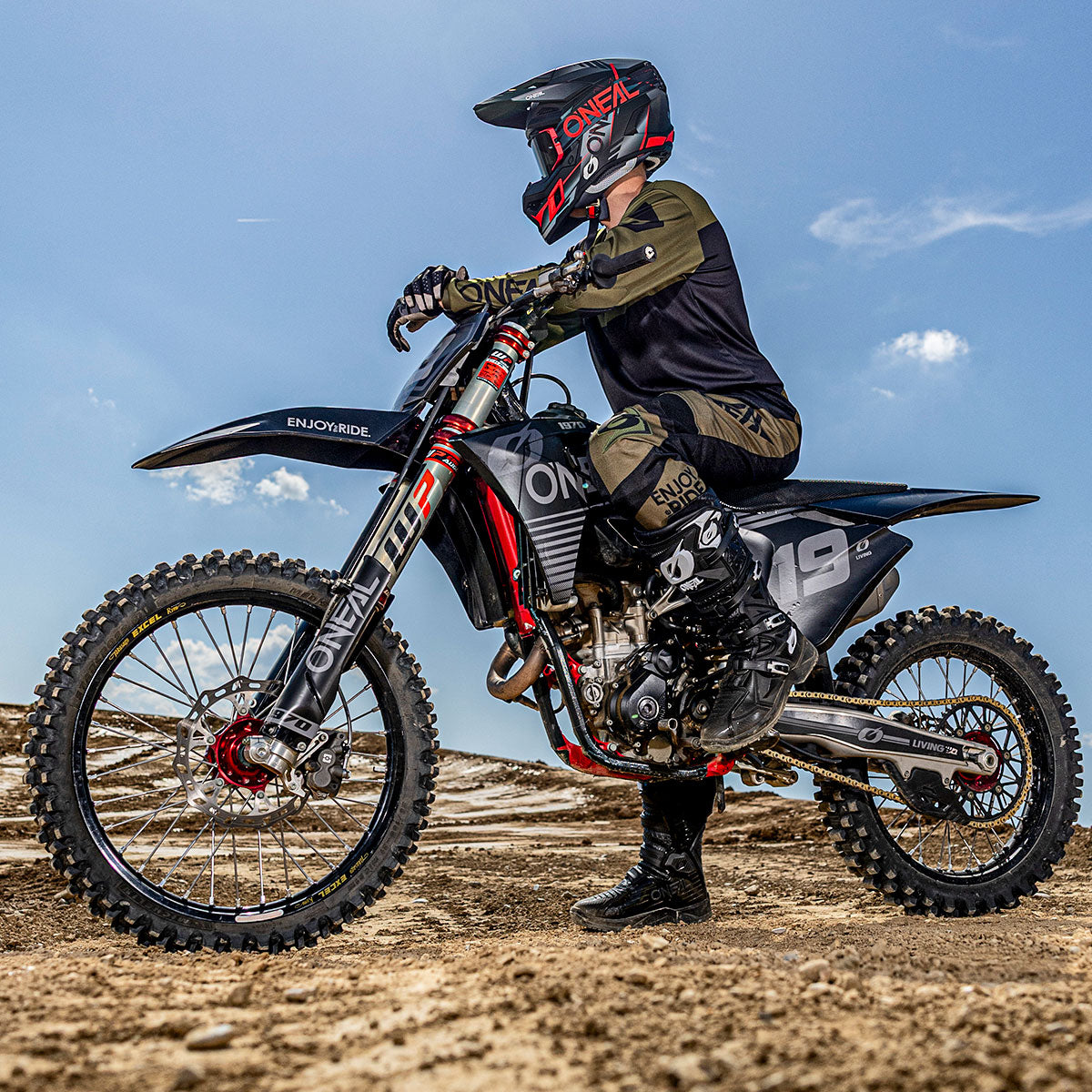 Eine Person in schwarzer Motocross-Kleidung und Helm sitzt auf einem schwarzen Dirt Bike mit dem Schriftzug "ONEAL", das auf einem felsigen Gelände unter einem klaren blauen Himmel geparkt ist.