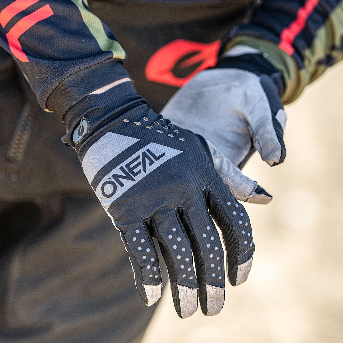 Eine Person, die schwarze und graue Fahrradhandschuhe der Marke O'NEAL trägt, stellt die Passform an einer Hand ein. Die Handschuhe haben weiße und graue Akzente mit kleinen Griffpunkten an den Fingern.