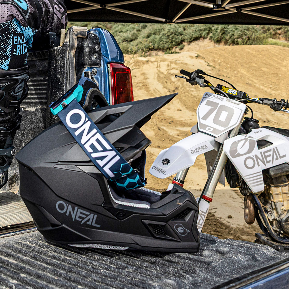 Ein schwarzer Motocross-Helm und eine Brille von O'Neal liegen auf einer Ladefläche eines Lastwagens, während im Hintergrund ein weißes Dirt Bike mit der Aufschrift "70" und "O'Neal" auf einer Sandpiste geparkt ist.