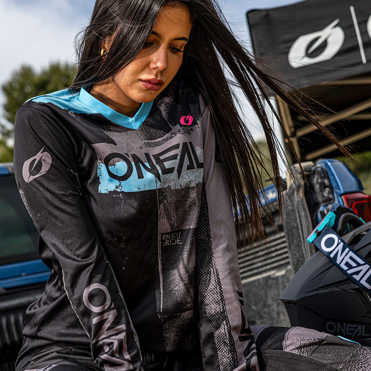 Eine Frau mit langen dunklen Haaren trägt ein schwarz-blaues O'Neal-Motocross-Trikot und steht im Freien in der Nähe von Fahrzeugen und einem Zelt, wobei neben ihr ein O'Neal-Helm zu sehen ist.