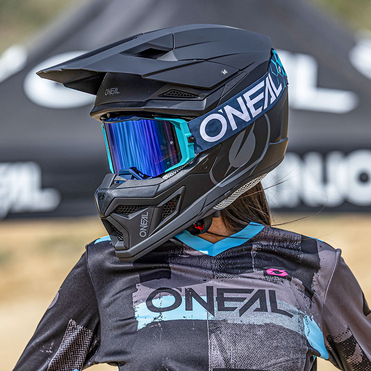 Eine Person, die einen schwarz-blauen O'NEAL-Motocross-Helm mit blauem Visier und ein passendes O'NEAL-Trikot trägt, steht im Freien und blickt zur Seite. Der Hintergrund ist unscharf und zeigt einen Teil eines Zeltes mit dem O'NEAL-Logo.