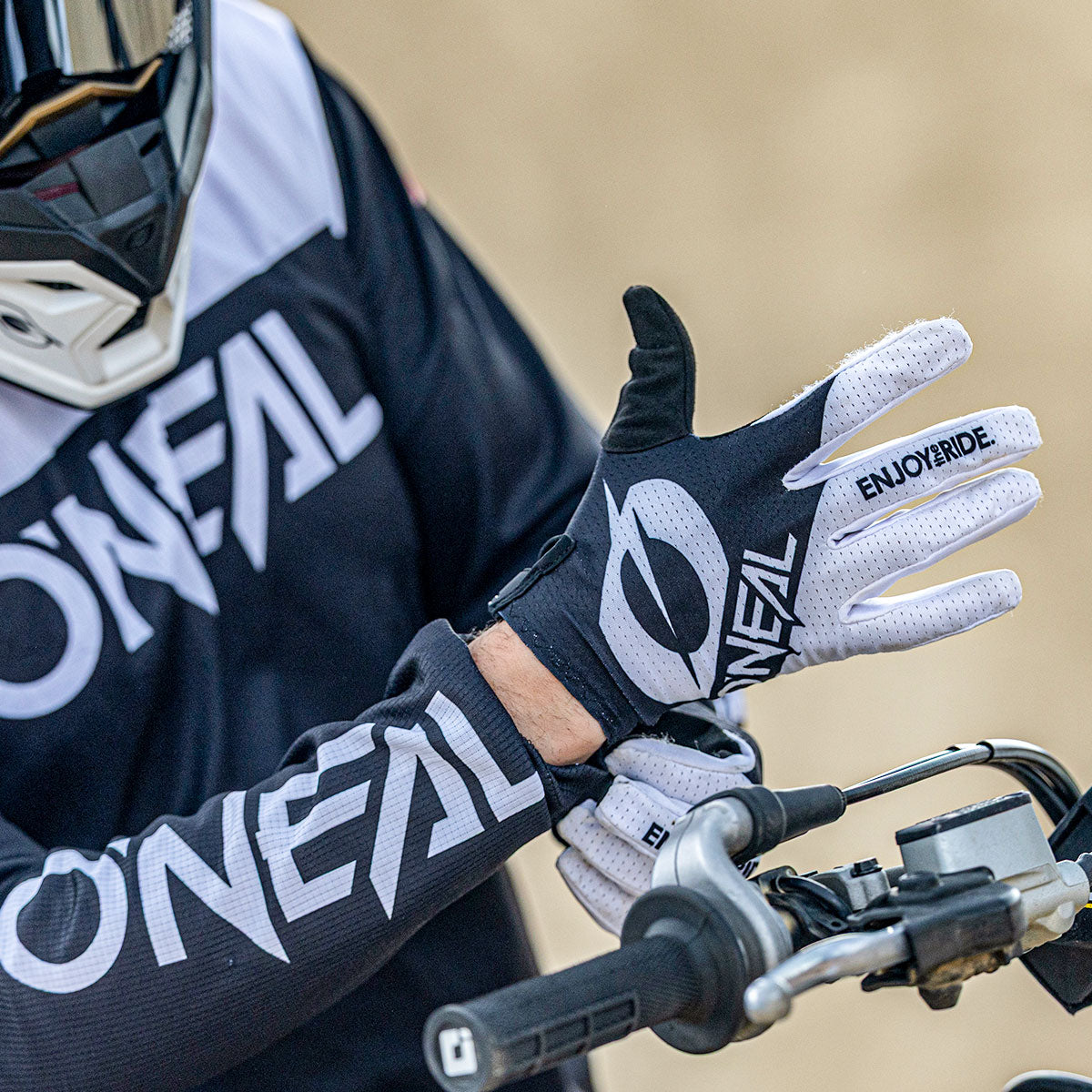 Eine Person, die ein schwarz-weißes O'NEAL-Motocross-Trikot und Handschuhe trägt, passt ihren Handschuh an, während sie den Lenker eines Motorrads hält. Das Wort "ENJOYRIDE" ist auf dem Handschuh aufgedruckt.