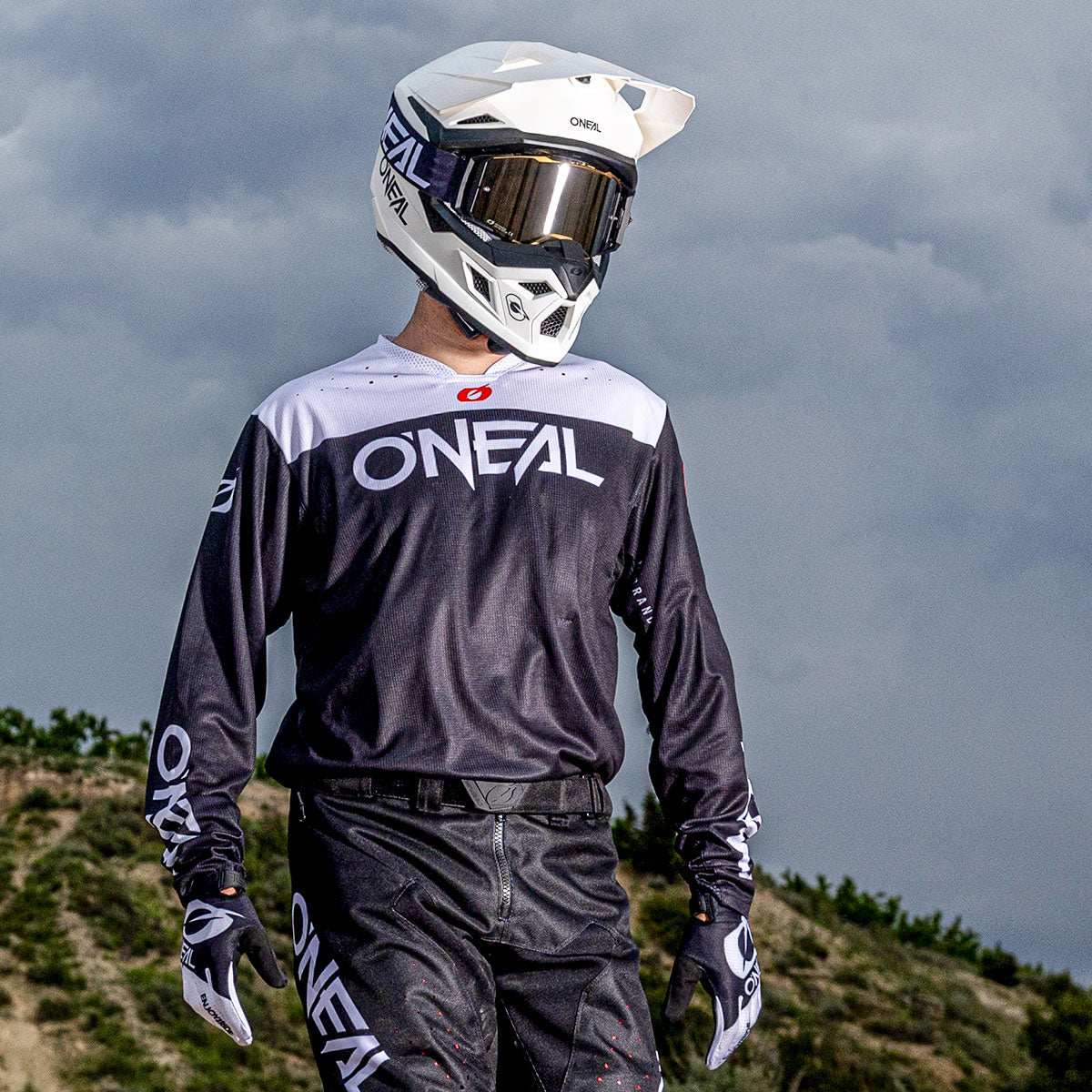 Ein Motocross-Fahrer in voller Montur, einschließlich weißem Helm, Schutzbrille, Handschuhen und schwarz-weißem O'NEAL-Trikot, steht im Freien vor einem wolkenverhangenen Himmel und grünen Hügeln im Hintergrund.