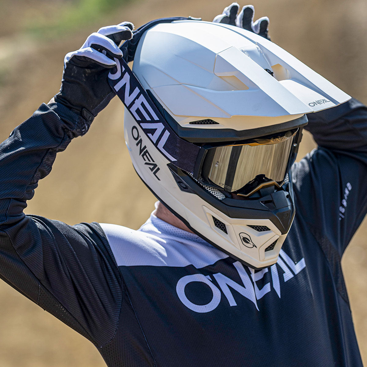 Eine Person mit schwarzen Handschuhen passt einen weißen O'NEAL-Motocross-Helm und eine Schutzbrille an. Sie ist mit einem passenden schwarz-weißen O'NEAL-Trikot bekleidet und steht im Freien auf einer unbefestigten Strecke.