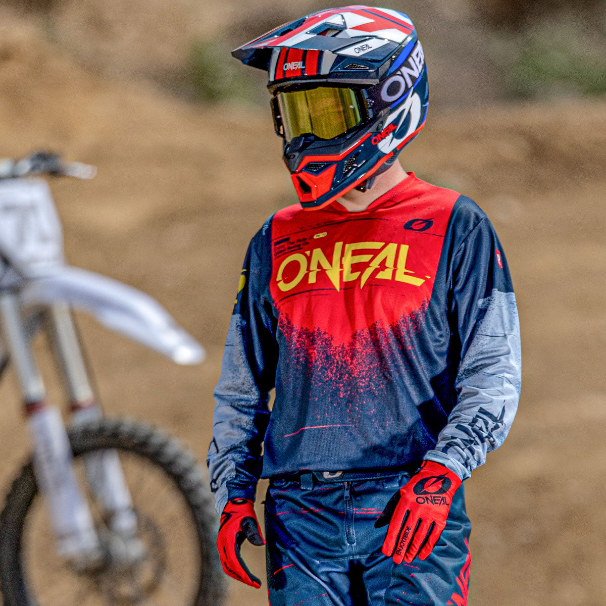 Eine Person, die ein komplettes Motocross-Outfit mit Helm, Handschuhen und Trikot mit "O'NEAL"-Logo trägt, steht im Freien neben einem unscharfen weißen Dirt Bike auf einer unbefestigten Strecke.