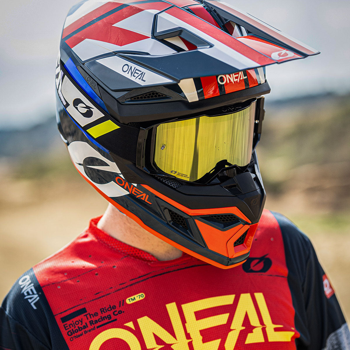 Eine Person, die einen bunten Motocross-Helm mit gelbem Visier und ein rot-schwarz-weißes O'Neal-Trikot trägt, steht im Freien vor einem unscharfen natürlichen Hintergrund.