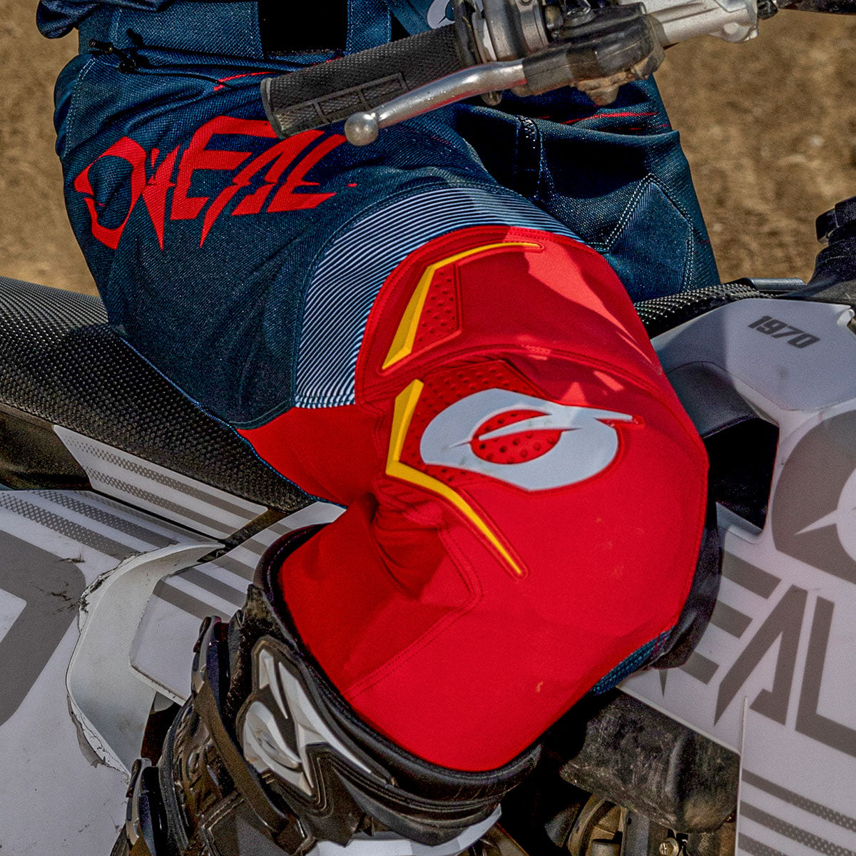 Nahaufnahme einer Person in rot-blauer Motorradkleidung und Knieschonern, die auf einem weiß-grauen Dirt Bike mit sichtbaren Markenlogos sitzt.