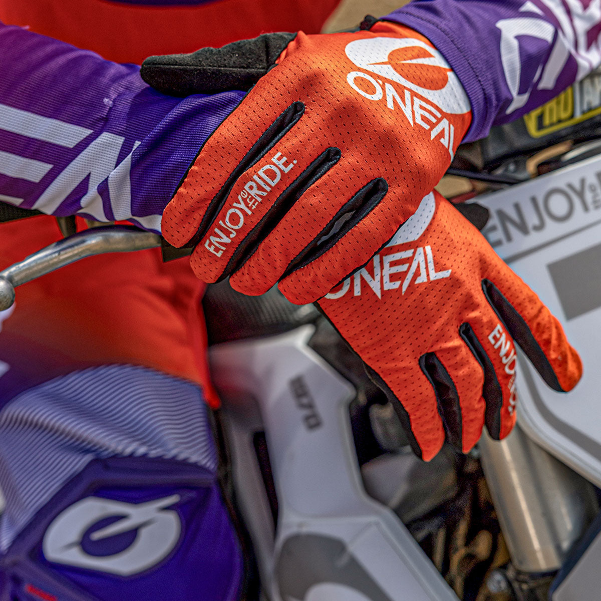 Nahaufnahme einer Person, die leuchtend orangefarbene O'Neal-Motocross-Handschuhe mit der Aufschrift "ENJOY THE RIDE" trägt und ihre Hände auf ein weißes Dirt Bike stützt. Die Person ist in lila und roter Kleidung gekleidet.