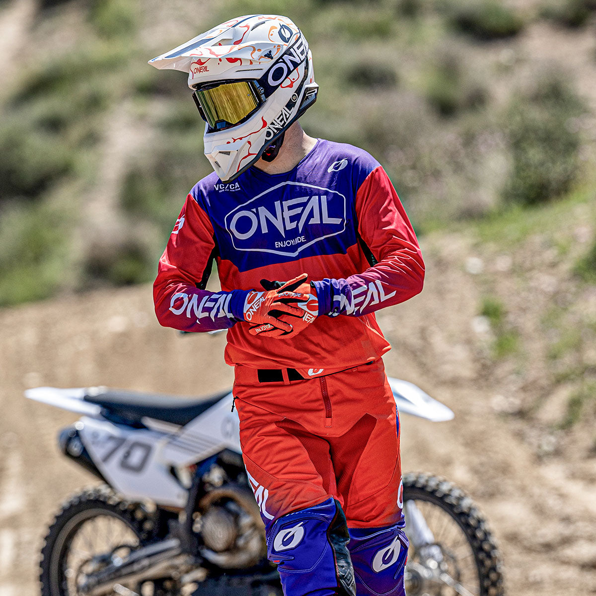 Ein Motocross-Fahrer in roter, blauer und lilafarbener O'Neal-Ausrüstung und weißem Helm steht auf einer unbefestigten Strecke mit einem weißen Dirt Bike im Hintergrund, umgeben von Grün.