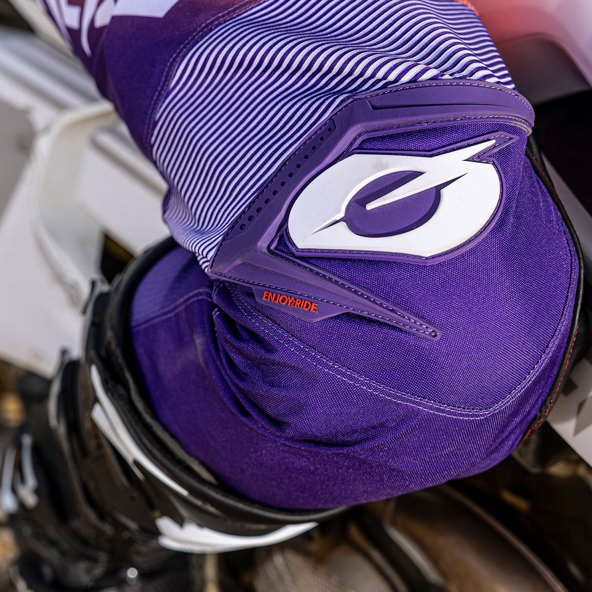 Nahaufnahme einer Person in lila-weißer Motocross-Kleidung mit einem stilisierten Buchstaben "O" und dem Wort "ENJOYRIDE" auf der Hose, die auf einem Motorrad sitzt.