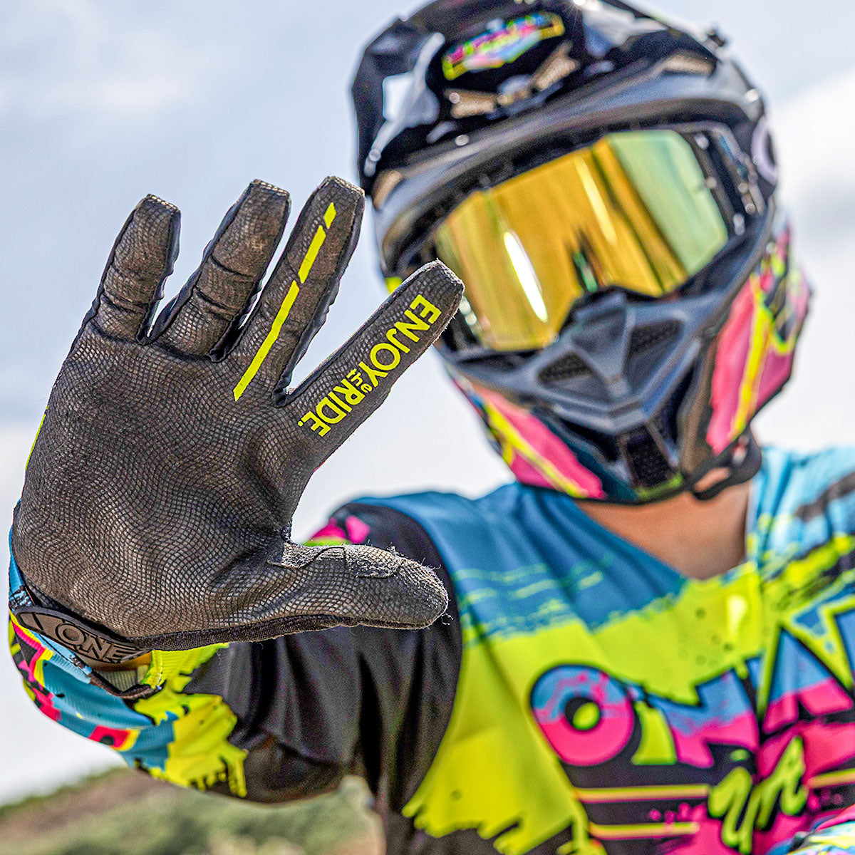 Eine Person in bunter Motocross-Kleidung und mit Helm hält eine behandschuhte Hand in Richtung Kamera. Auf dem Handschuh steht "ENJOY RIDE" und die Person trägt eine reflektierende Schutzbrille vor einem unscharfen Hintergrund.
