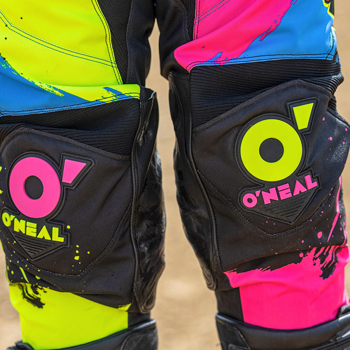 Nahaufnahme einer knallbunten Motocross-Hose mit neongelben, pinken und grünen Mustern und dem O'Neal-Logo auf jedem Kniepolster. Die Hose wird von einer Person getragen, die auf sandigem Boden steht.