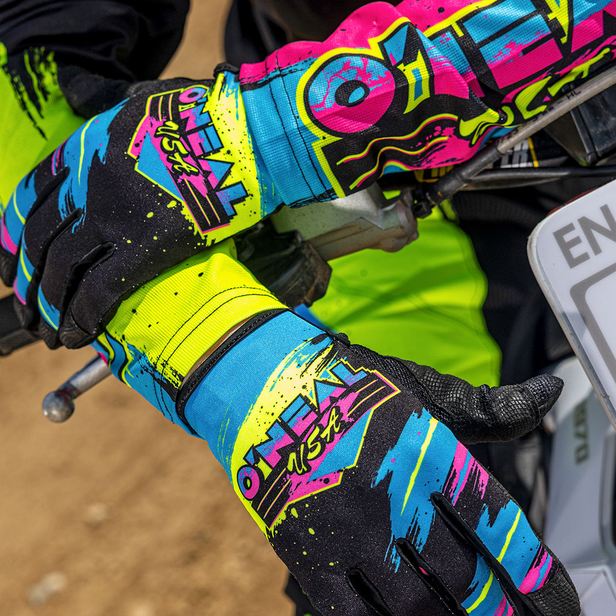 Nahaufnahme einer Person mit bunten, gemusterten Motocross-Handschuhen von O'Neal USA, die den Lenker eines Dirt-Bikes hält. Der Hintergrund zeigt eine Dirt Track.