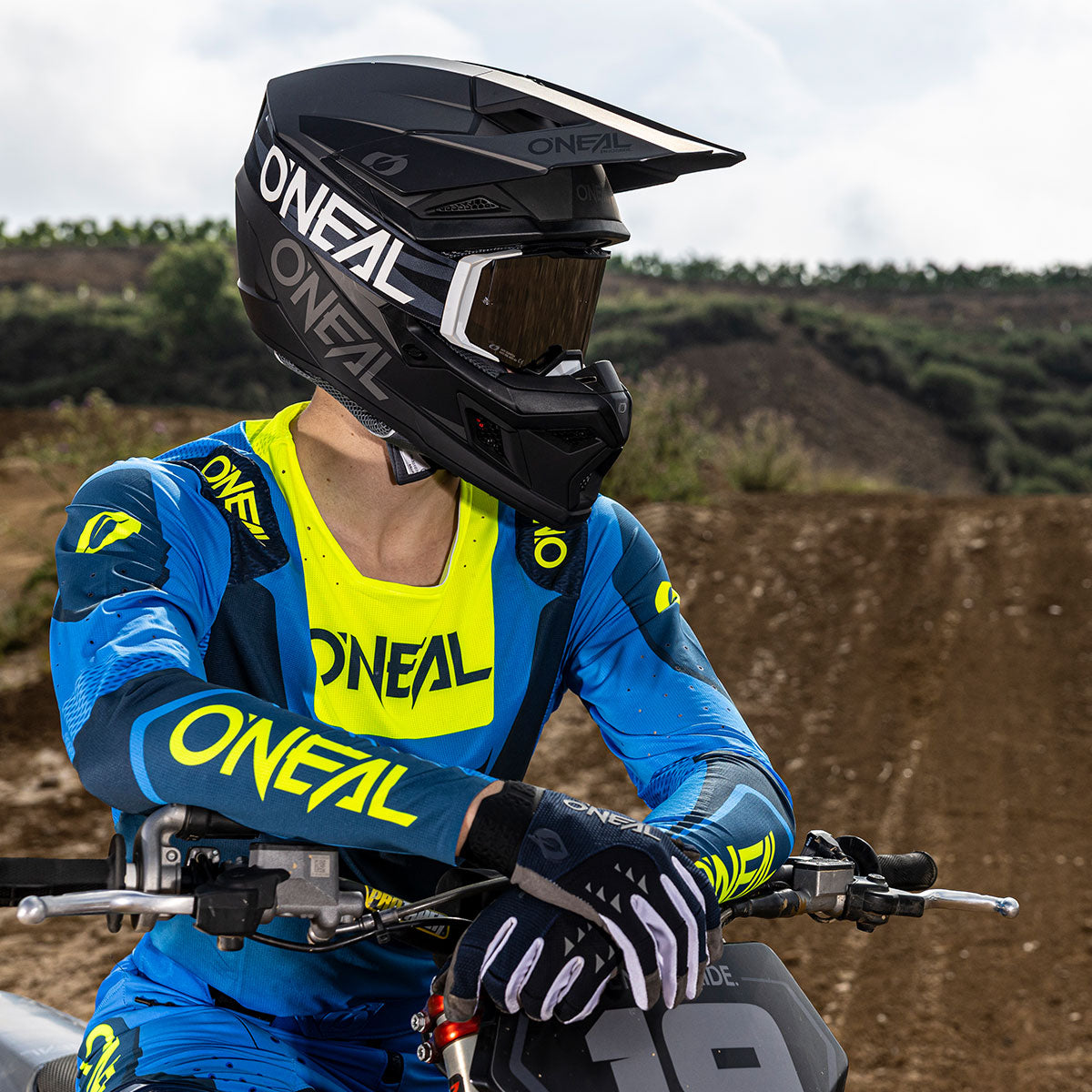 Ein Motocross-Fahrer in blau-gelber O'Neal-Kleidung und schwarzem Helm sitzt auf einem Geländemotorrad und stützt sich mit den Armen auf den Lenker, während im Hintergrund eine unbefestigte Strecke und Grünflächen zu sehen sind.