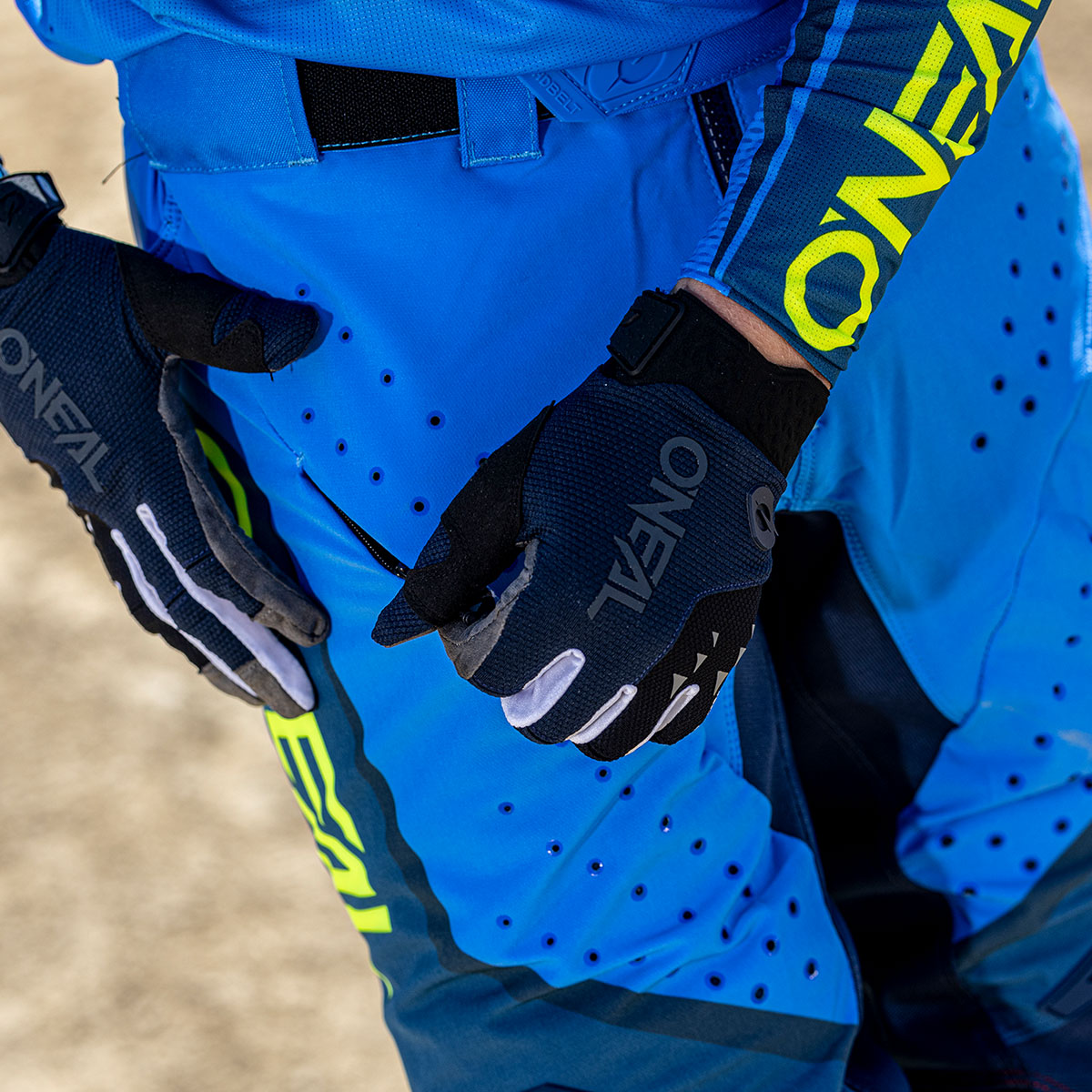 Eine Person, die eine blau-schwarze O'NEAL-Motocross-Ausrüstung und Handschuhe trägt, zieht den Reißverschluss ihrer Hose zu. Die Ausrüstung weist gelbe Akzente und ein Branding auf, mit einem sandigen Boden im Hintergrund.