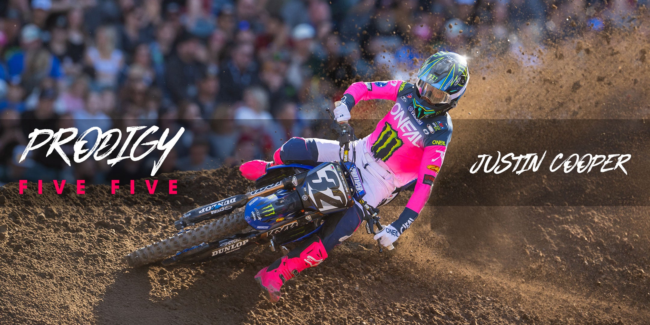 Ein Motocross-Fahrer in pink-blauer Ausrüstung rast über eine unbefestigte Strecke und wirbelt dabei Staub auf. Die Textüberlagerung lautet „Prodigy Five Five“ und „Justin Cooper“. Im unscharfen Hintergrund ist eine Menschenmenge zu sehen.