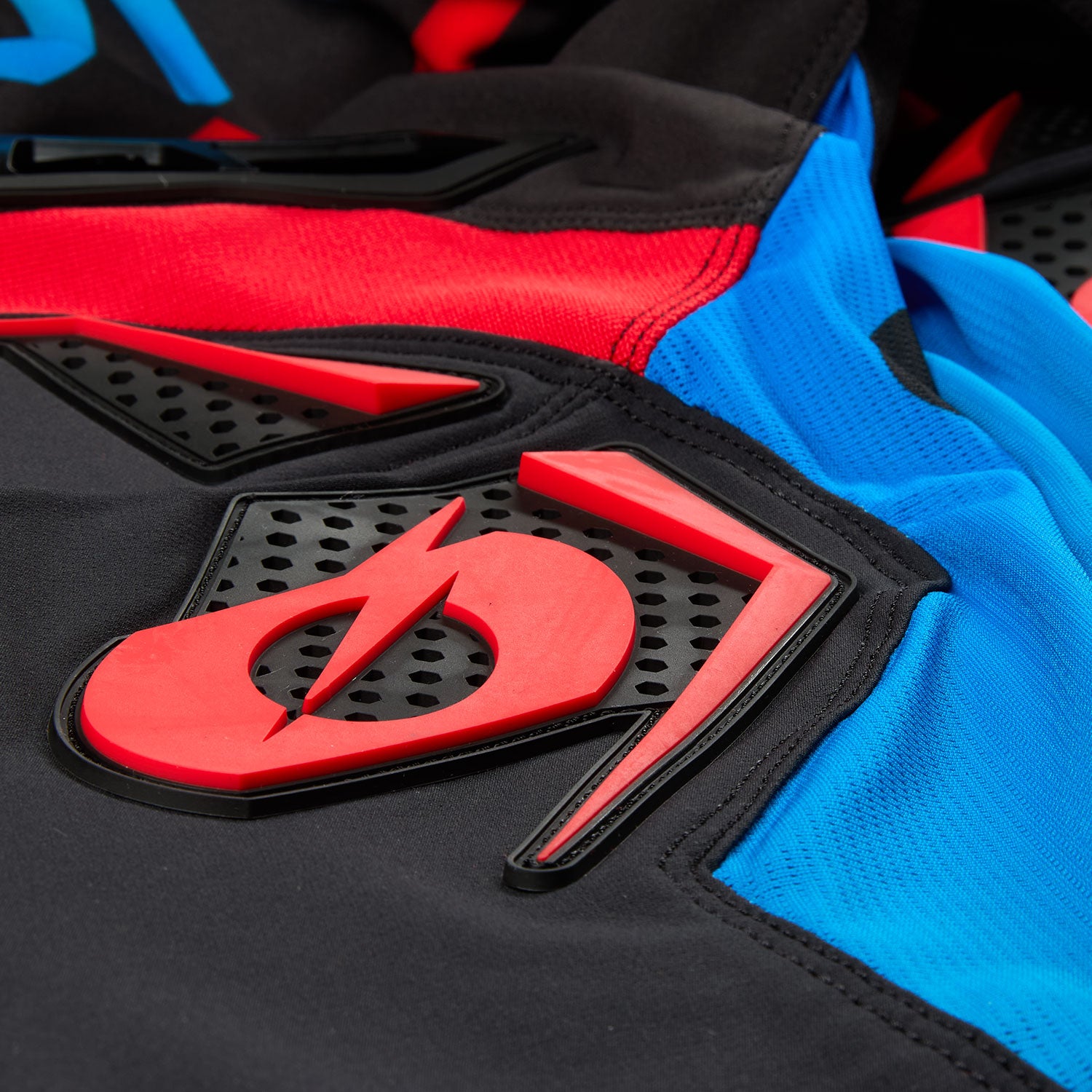Nahaufnahme eines Stoffes mit einem roten Blitz-Logo, das in einem Kreis mit einer schrägen Linie eingeschlossen ist, vor schwarzen, roten und blauen Abschnitten. Das Design umfasst sowohl strukturierte als auch glatte Oberflächen und zeigt komplizierte Nähte und langlebige Materialien.