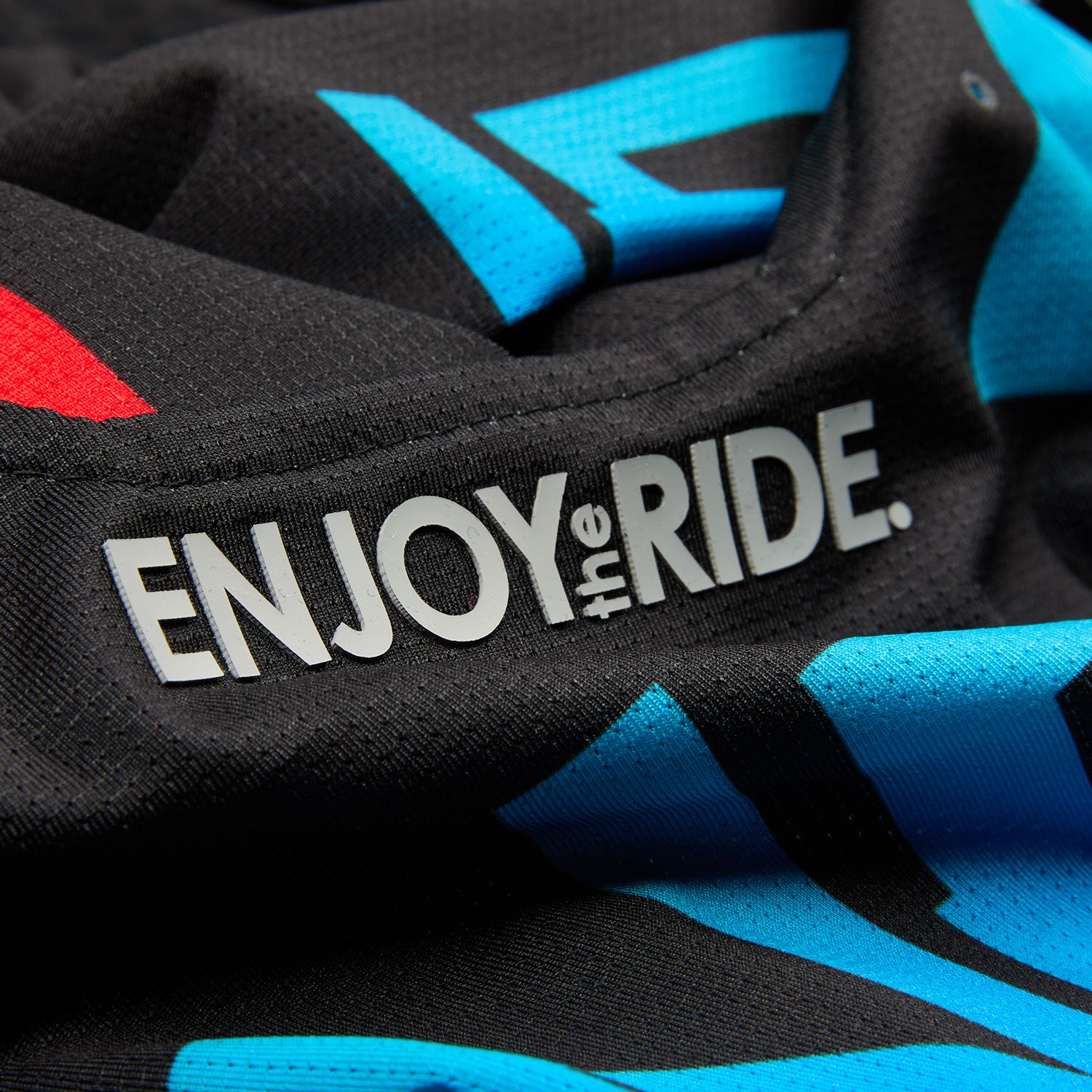 Nahaufnahme eines schwarzen Stoffes mit dem in Weiß gedruckten Text „ENJOY the RIDE“. Der Stoff scheint Teil eines Sporttrikots oder eines ähnlichen Sportbekleidungsstücks zu sein und weist zusätzliche blaue und rote Designelemente auf dem schwarzen Hintergrund auf.