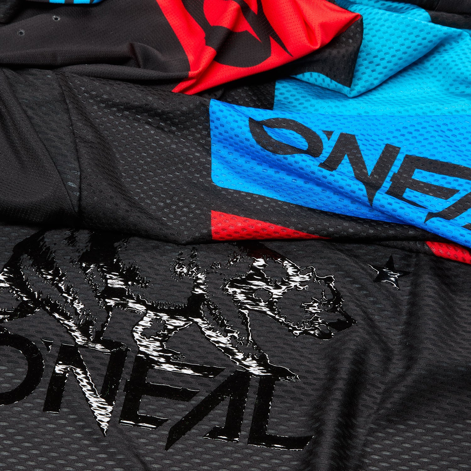 Nahaufnahme eines Stoffes mit dem O'Neal-Markenlogo in Hellblau und einer stilisierten Bärengrafik sowie roten und blauen geometrischen Elementen auf einem schwarzen strukturierten Hintergrund.