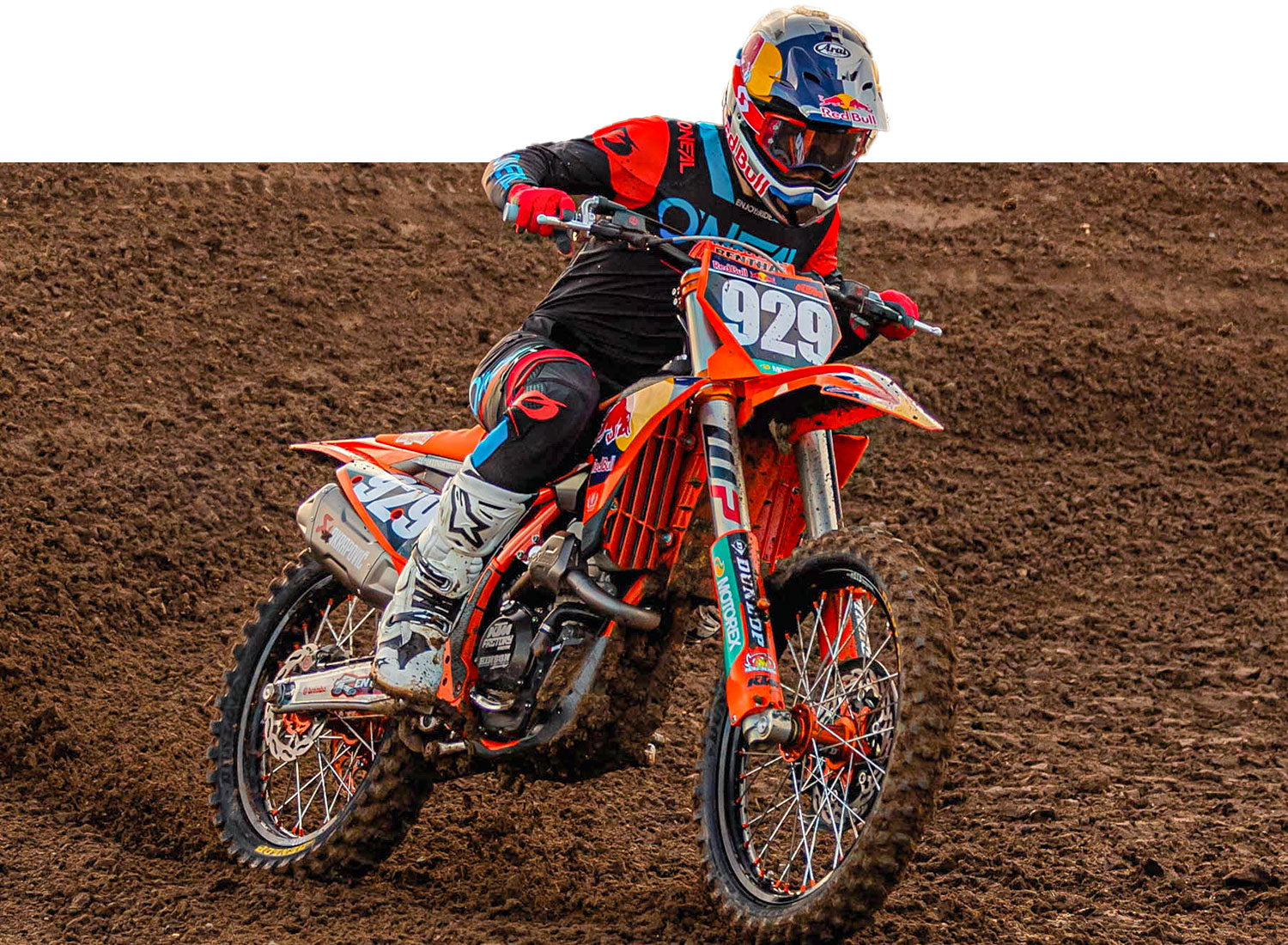Ein Motocrossfahrer in rot-schwarzem Outfit, Helm und Schutzbrille fährt ein orangefarbenes Dirtbike mit der Nummer 929 auf der Vorderseite. Der Fahrer fährt auf einer unbefestigten Strecke mit sichtbaren Reifenprofilen und Schlamm. Der Hintergrund ist größtenteils klar und hebt den Fahrer und das Motorrad hervor.