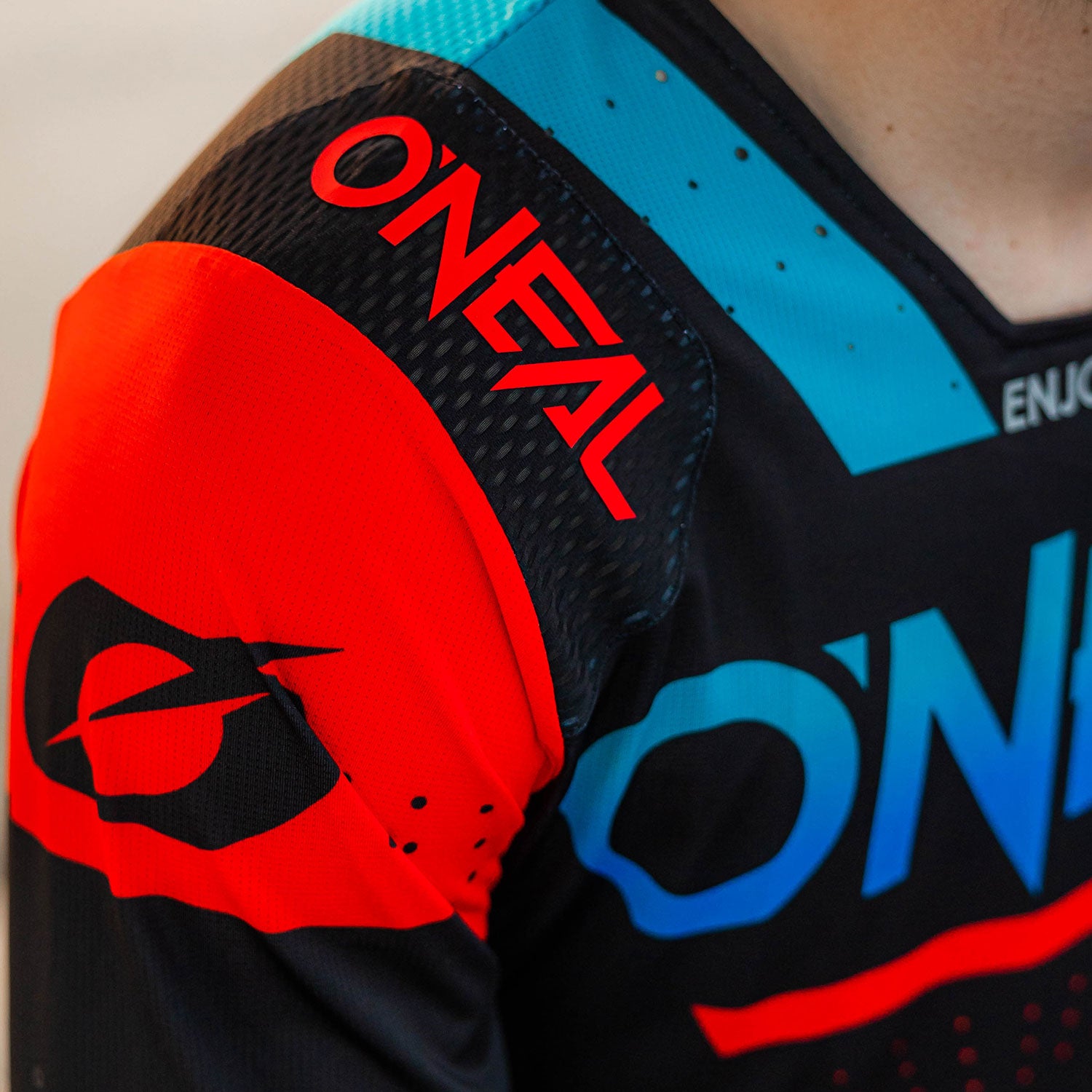 Nahaufnahme einer Person, die ein farbenfrohes Motocross-Trikot trägt, das hauptsächlich die Marke O'NEAL zeigt. Das Trikot hat eine Farbmischung aus Schwarz, Rot und Blau, mit Logos und grafischen Designs im Schulter- und Brustbereich.