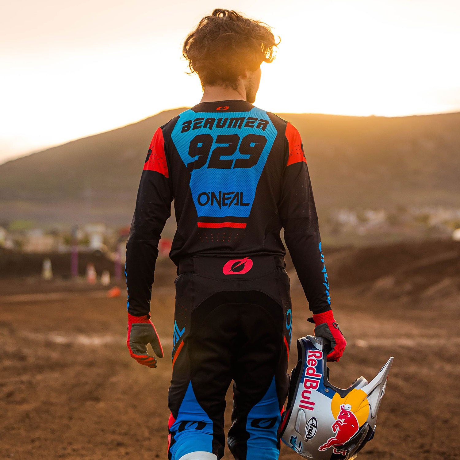 Ein Motocrossfahrer steht auf einer Dirt-Piste und hält einen Helm mit Red Bull-Logo in der Hand. Auf der Ausrüstung des Fahrers sind in Blau und Rot der Name „Breakum“ und die Nummer „929“ zu sehen. Im Hintergrund geht die Sonne hinter einer Hügellandschaft unter.