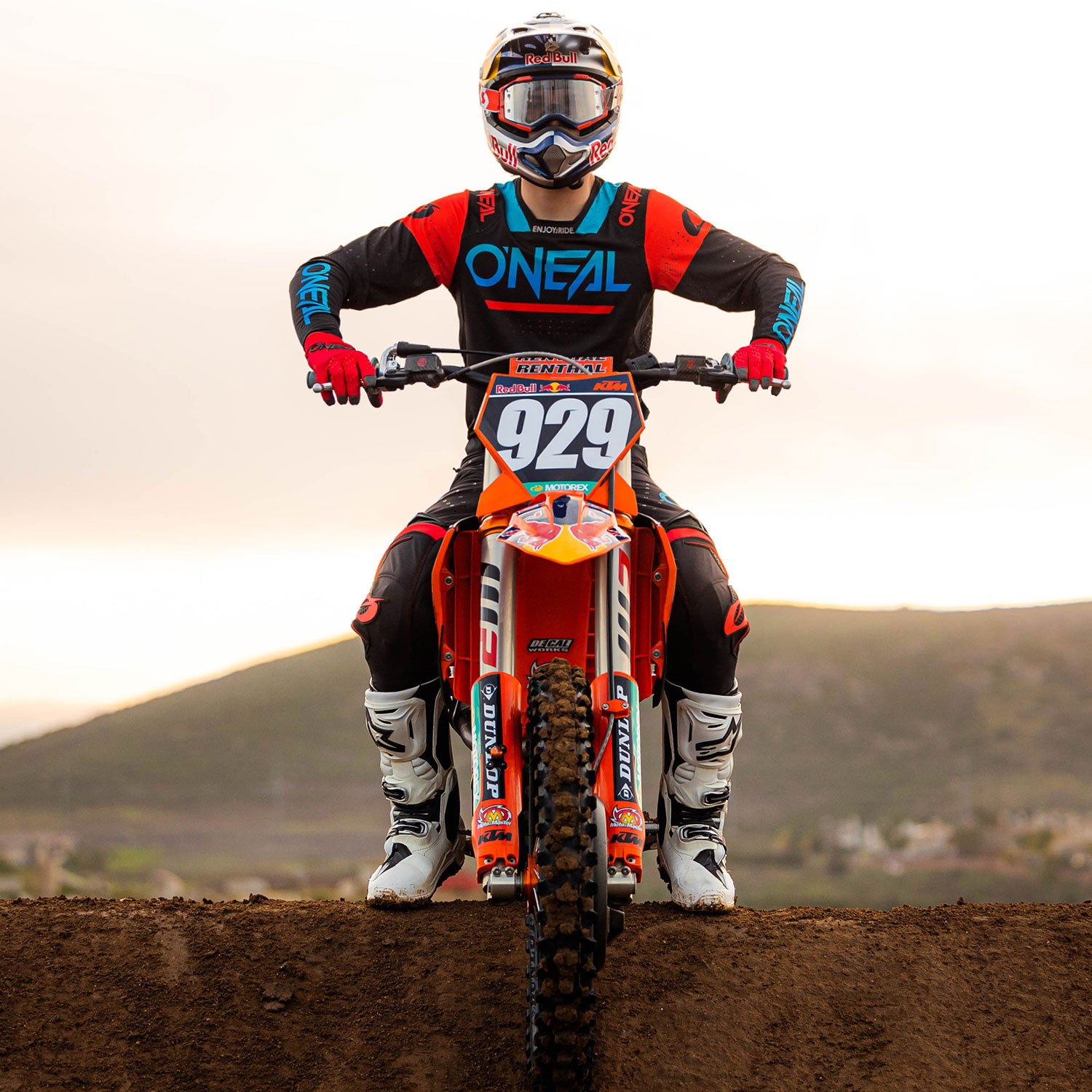 Ein Motocross-Fahrer in voller Montur, einschließlich Helm, Schutzbrille und rot-schwarzem Outfit, sitzt auf einem roten Motocross-Motorrad mit der Nummer 929. Sie stehen auf einer unbefestigten Strecke mit einer hügeligen Landschaft im Hintergrund unter einem bewölkten Himmel.