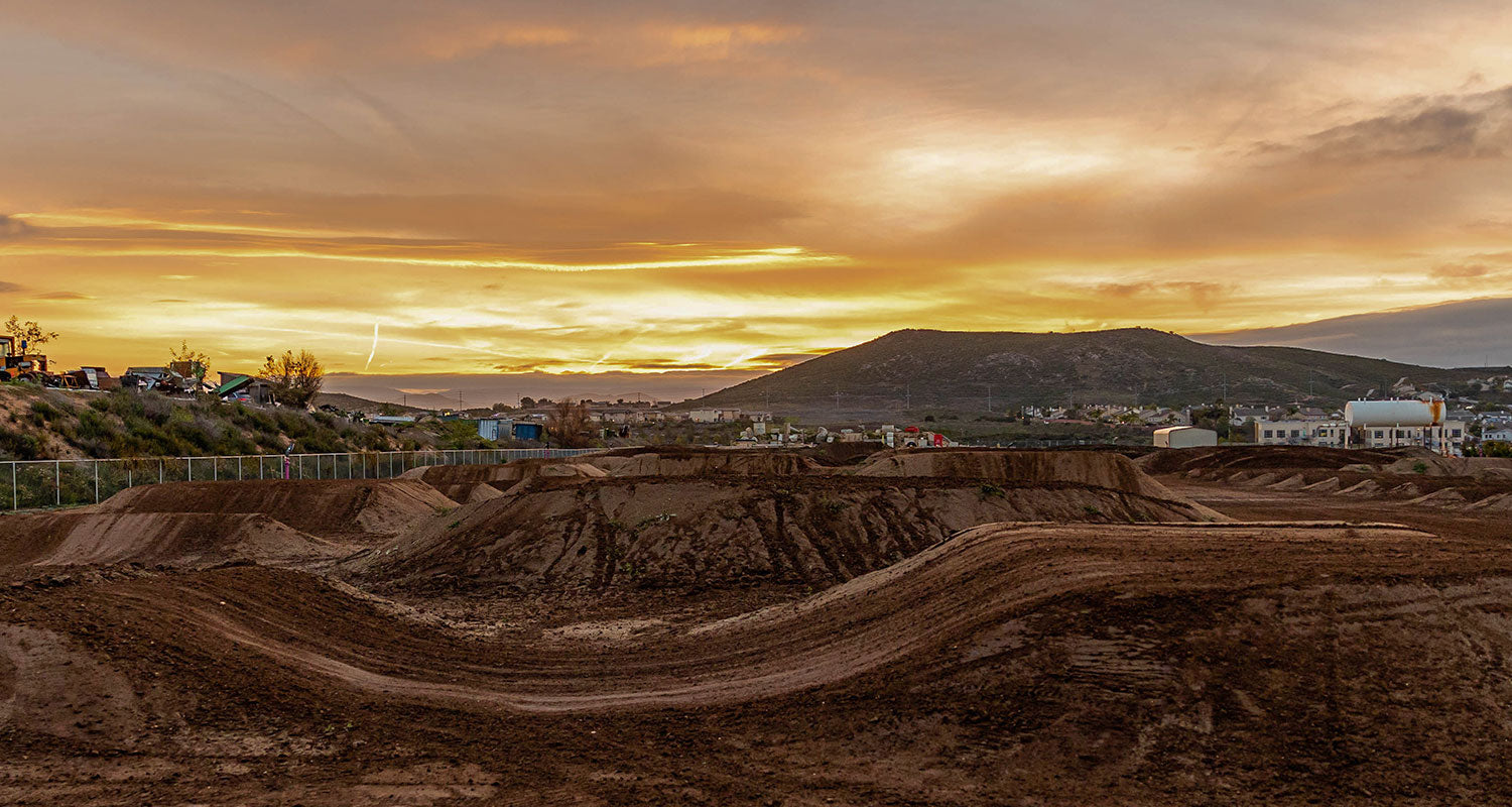 Das Bild zeigt einen Sonnenuntergang über einer Dirtbike-Strecke mit welligen Hügeln und Sprüngen. Im Hintergrund sind entfernte Gebäude und Berge unter einem Himmel in goldenen und orangen Farbtönen zu sehen. Das Gebiet ist von etwas Grün und einem Zaun umgeben.