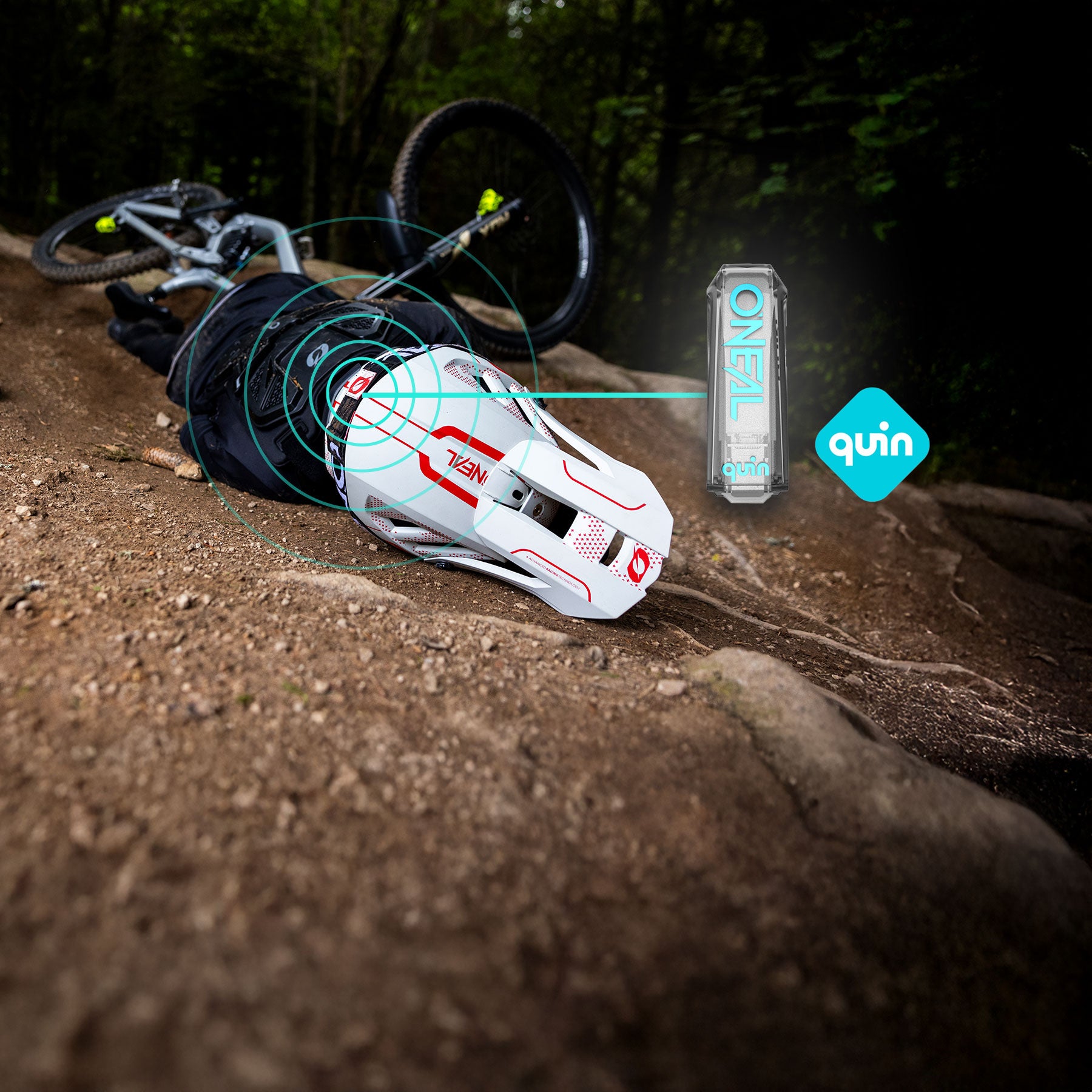 Ein Radfahrer liegt nach einem Sturz auf einem Waldweg. Er trägt einen weißen Helm, von dem blaue digitale Signale ausgehen, die auf eine vernetzte Sicherheitsfunktion hinweisen. In der Nähe befinden sich eine Markendose und das Quin-Logo.
