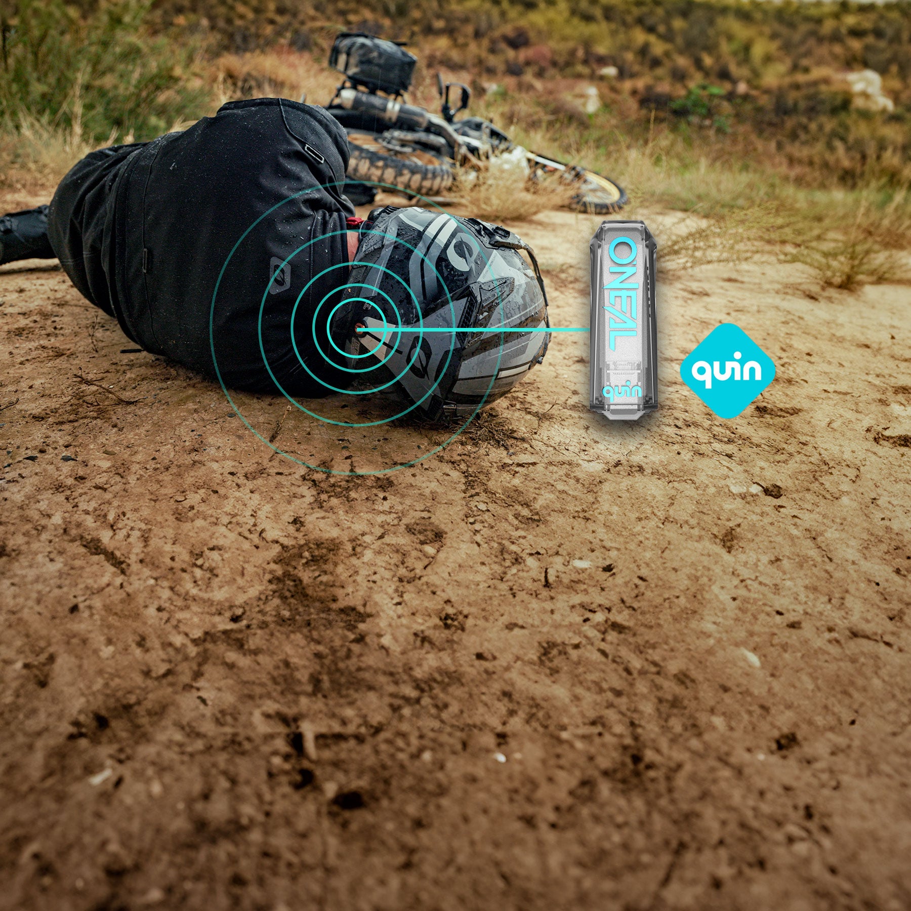 Ein Mountainbiker liegt nach einem Sturz auf einem Feldweg, daneben ein Fahrrad. Der Biker trägt Helm und Ausrüstung. Digitale Grafiken heben den Helm hervor und zeigen das Quin-Logo und ein ONETAL-Gerät.