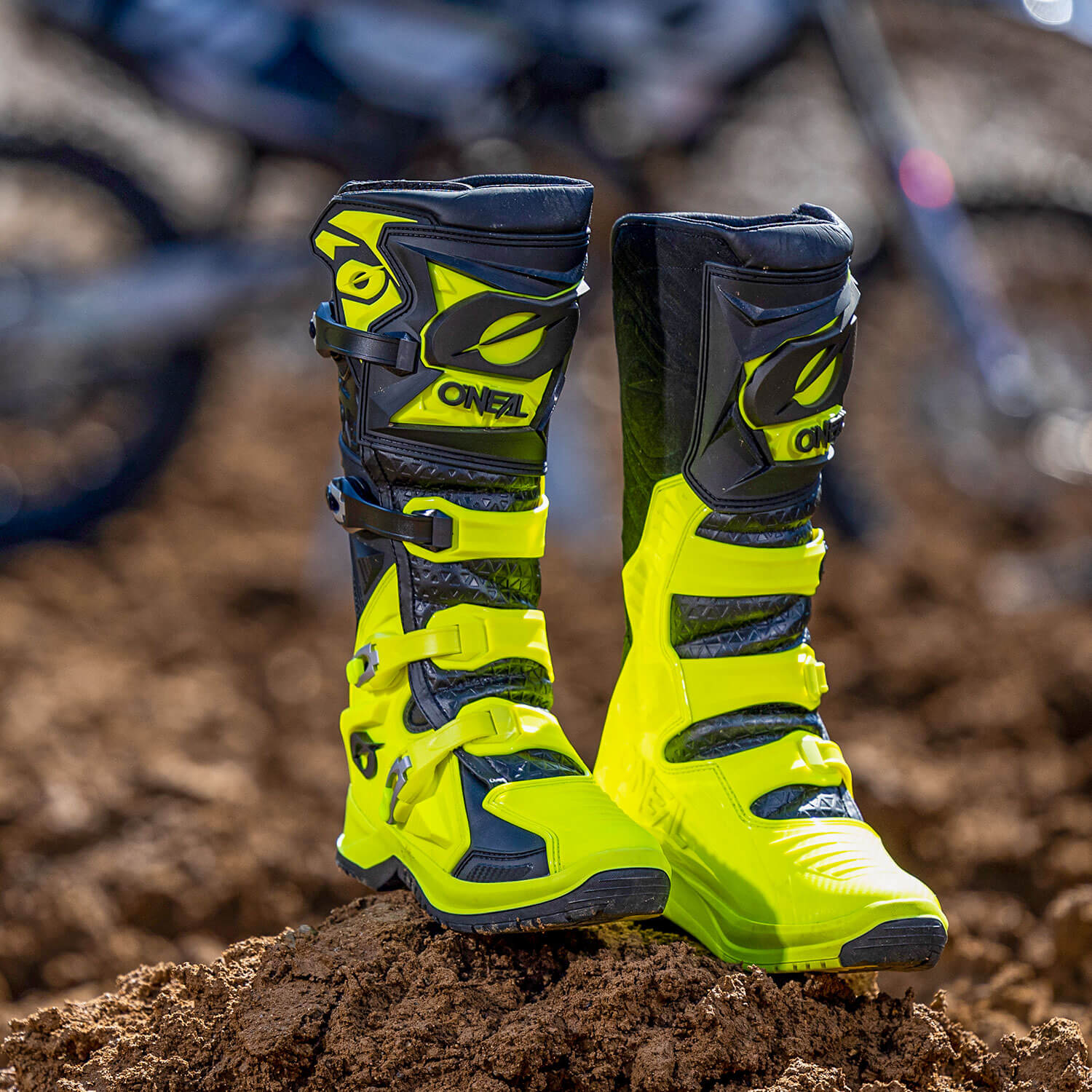 Ein Paar neongelbe und schwarze Motocross-Stiefel mit mehreren Schnallen steht aufrecht auf einem Erdhaufen. Der Hintergrund ist leicht verschwommen und zeigt ein Motocross-Motorrad, was auf eine Offroad-Umgebung hindeutet.