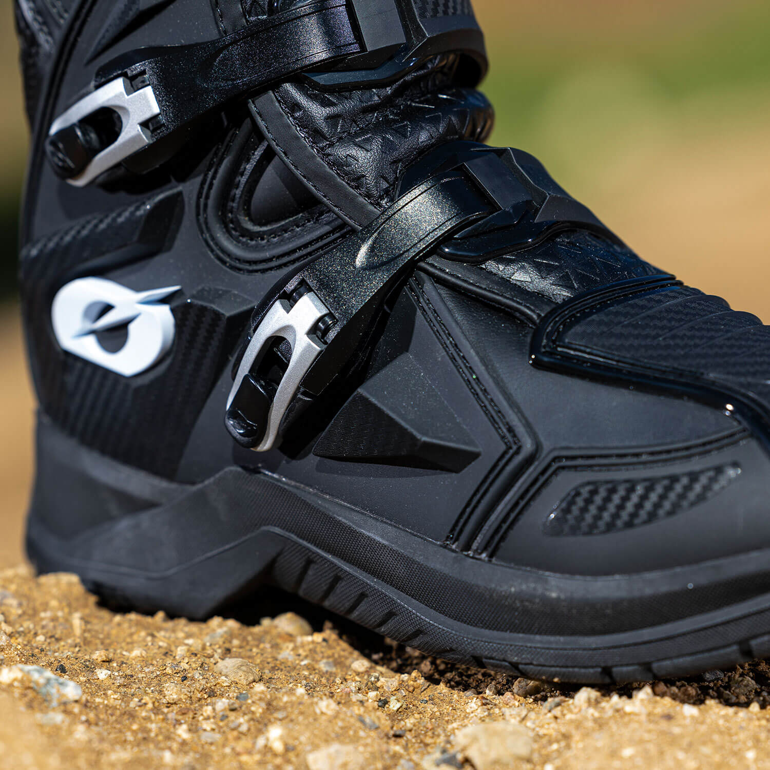 Nahaufnahme eines schwarzen Motocross-Stiefels auf einer unbefestigten Oberfläche. Der Stiefel hat silberne Schnallen, ein robustes Profil und ein Logo mit einem stilisierten Buchstaben „O“. Der Fokus liegt auf dem Mittelteil des Stiefels, wodurch sein Design und sein Befestigungssystem hervorgehoben werden.
