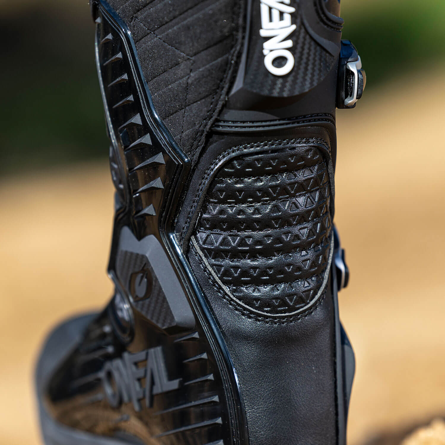 Nahaufnahme eines schwarzen Motocross-Stiefels mit detaillierten Nähten und strukturierten Mustern. Der Markenname „ONEAL“ ist deutlich auf dem Stiefel zu sehen und er hat Metallschnallen, um die Passform zu gewährleisten. Der Hintergrund ist eine verschwommene Außenkulisse mit einem Hauch von Grün.