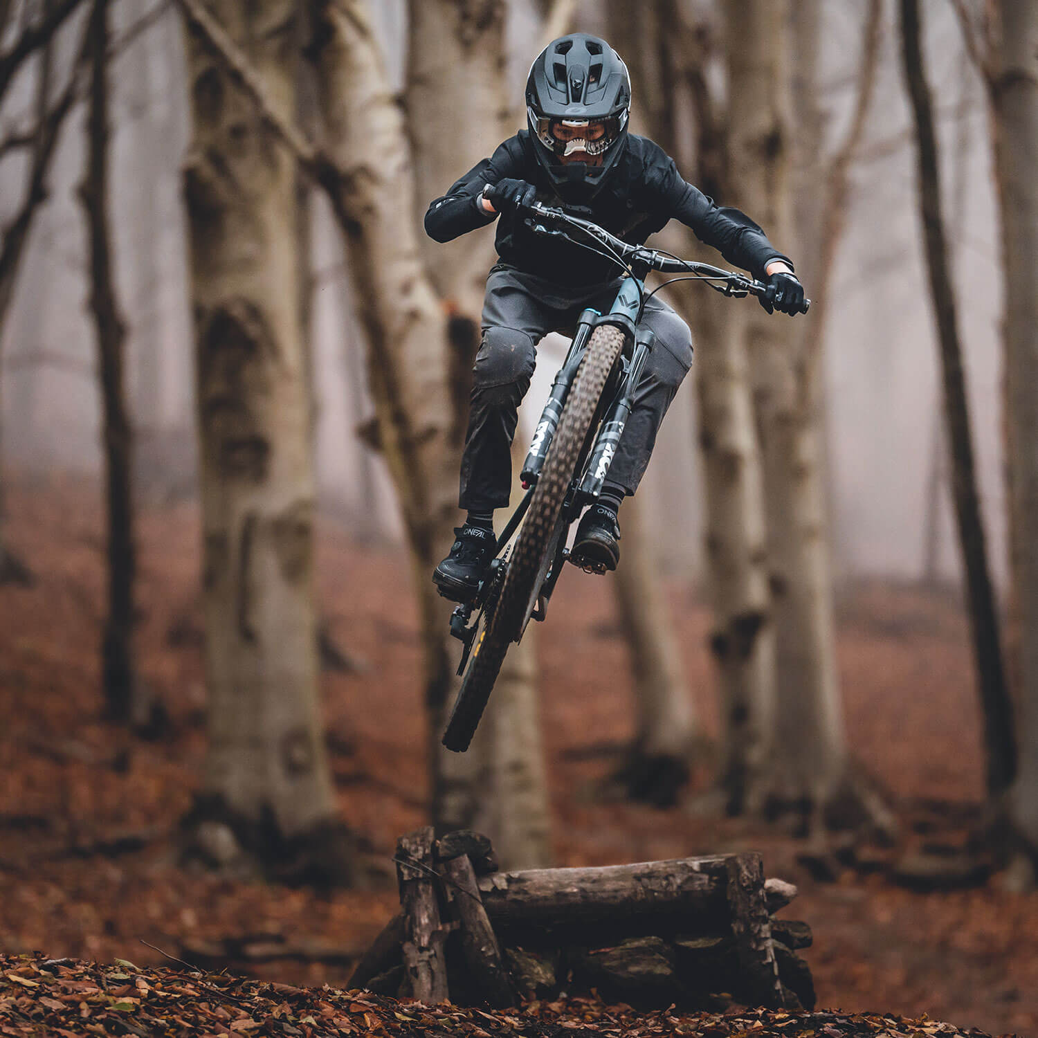 Eine Person mit Helm und Schutzausrüstung fährt Mountainbike und fängt einen Sprung in der Luft in einem nebligen Herbstwald mit hohen, blattlosen Bäumen im Hintergrund ein. Der Boden ist mit abgefallenen Blättern bedeckt, die zur Waldlandschaft beitragen.