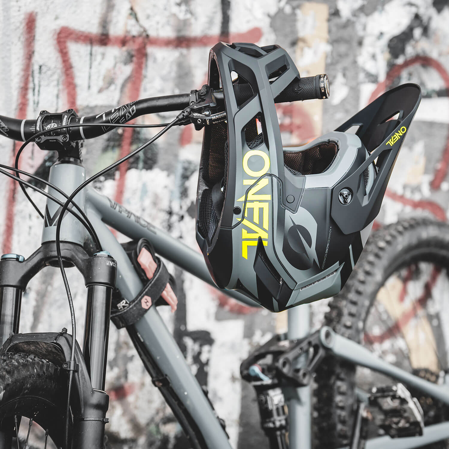 Ein Integralhelm mit der Marke „O'Neal“ hängt am Lenker eines grauen Mountainbikes. Die Szene hat einen urbanen Hintergrund mit Graffiti an einer Wand. Das Fahrrad verfügt über eine Vorderradgabel mit Federung und Stollenreifen, was darauf hindeutet, dass es für das Radfahren im Gelände konzipiert ist.