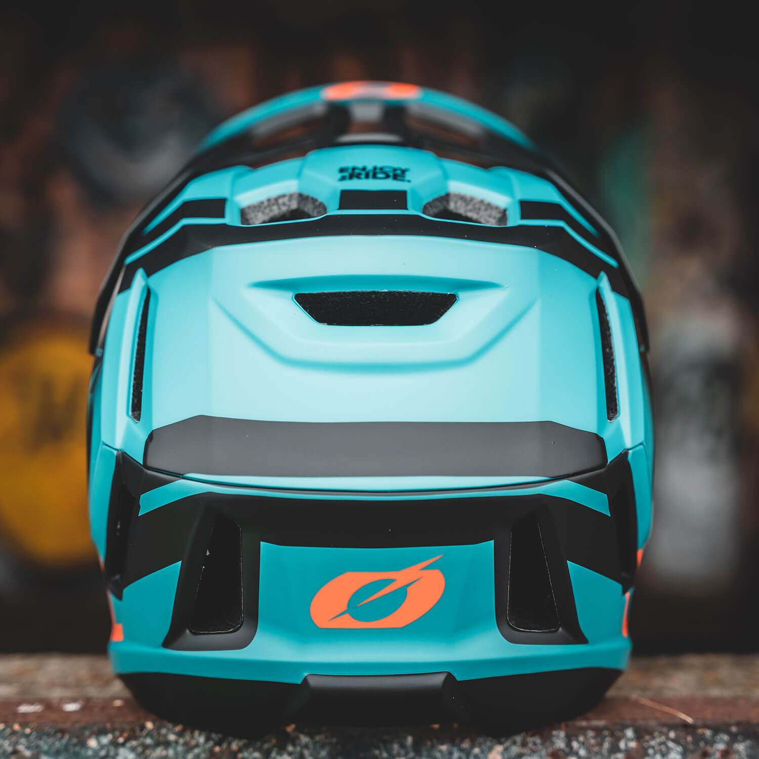 Eine Nahaufnahme der Rückseite eines cyan-schwarzen Fahrradhelms mit einem orangefarbenen Logo. Der Helm verfügt über mehrere Belüftungsöffnungen, ein stromlinienförmiges Design und ein kleines Visier im oberen Teil. Der unscharfe Hintergrund zeigt Gelb- und Brauntöne.