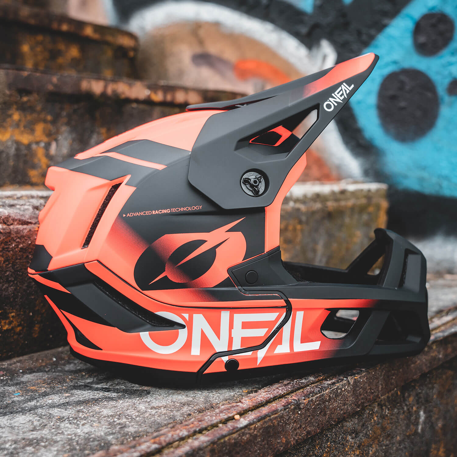 Ein farbenfroher O'Neal-Integralhelm für Mountainbikes steht auf einer Betontreppe im Freien, im Hintergrund ist Graffiti zu sehen. Der Helm ist rot-schwarz gestaltet und das Logo der Marke O'Neal ist deutlich zu sehen.