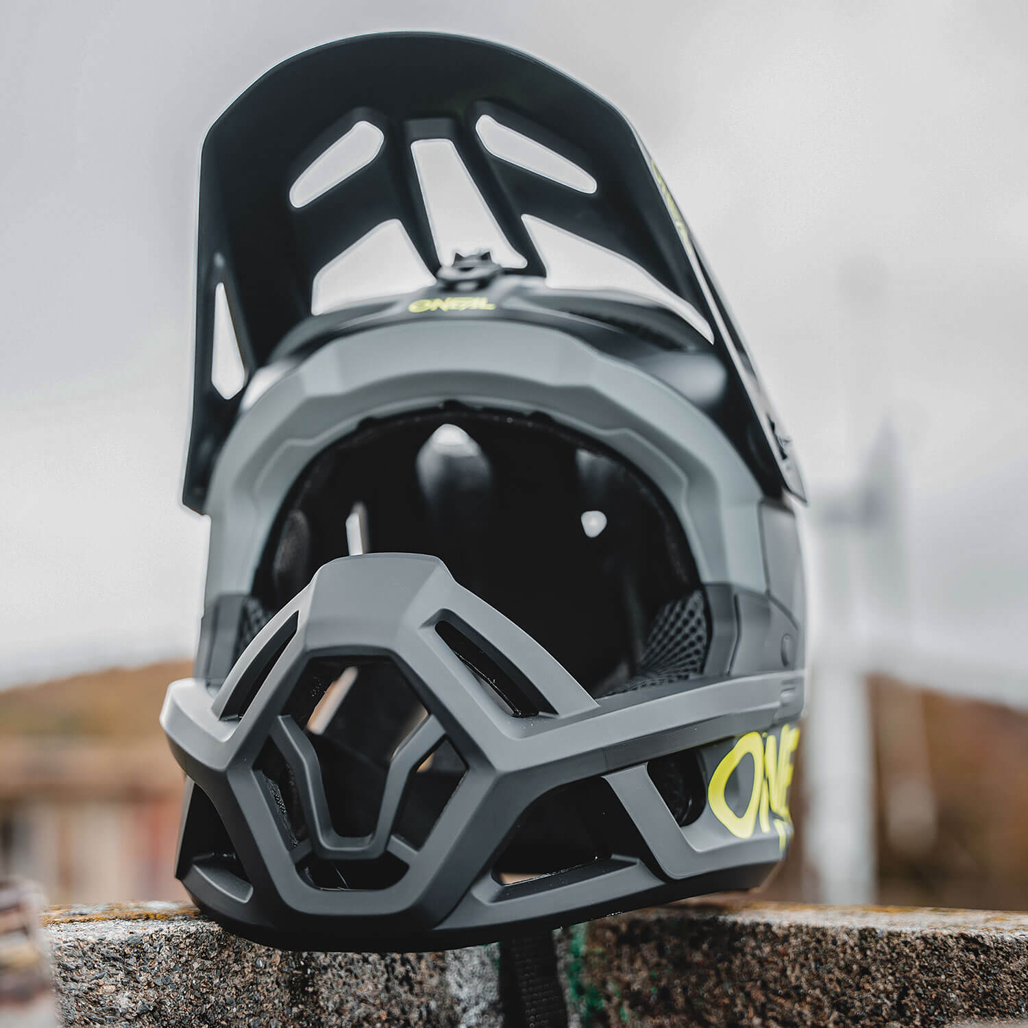 Nahaufnahme eines schwarz-grauen Mountainbikehelms mit Visier, der das ganze Gesicht schützt. Der Helm sieht robust aus und liegt auf einer Steinoberfläche vor einem verschwommenen Außenhintergrund.