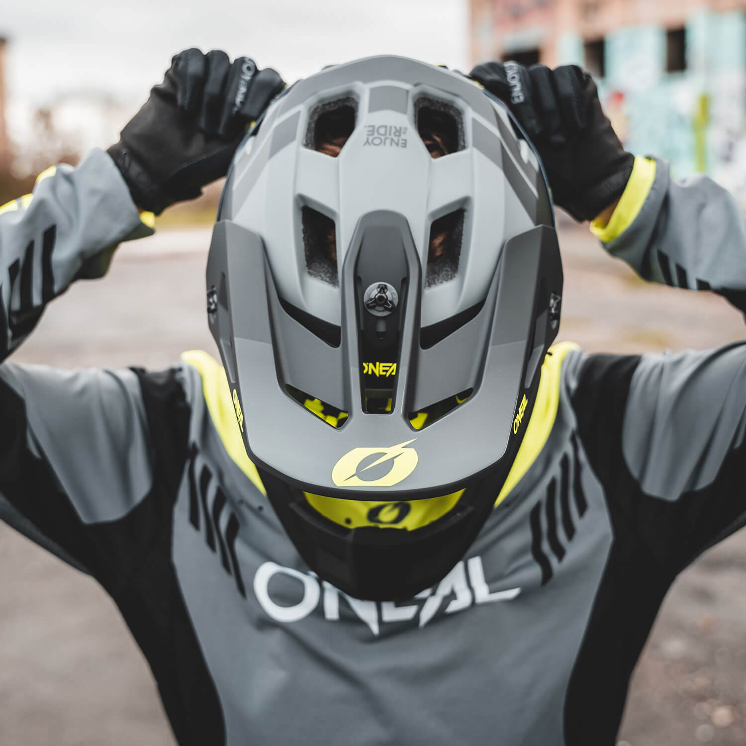 Eine Person mit einem grau-schwarzen Motocross-Helm mit gelben Akzenten und einem passenden Outfit mit der Marke „O'NEAL“ steht im Freien. Die Person rückt den Helm mit beiden Händen zurecht und bereitet sich auf eine Fahrt vor. Der Hintergrund ist leicht verschwommen, was auf eine städtische Umgebung hindeutet.