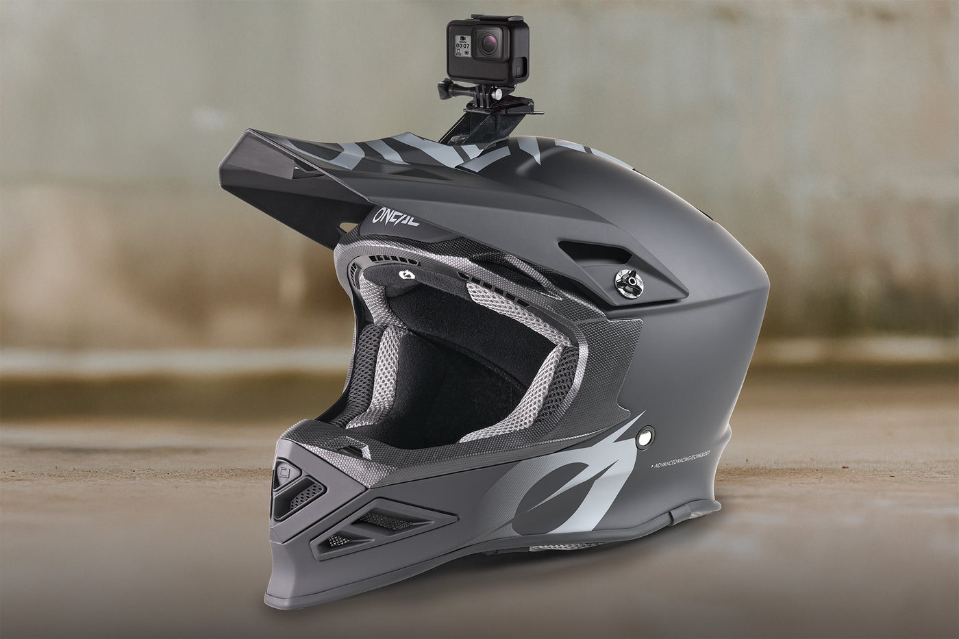 Ein mattgrauer Integralhelm für Motocross mit einer darauf montierten Action-Kamera, abgebildet vor einem unscharfen, neutralen Hintergrund.