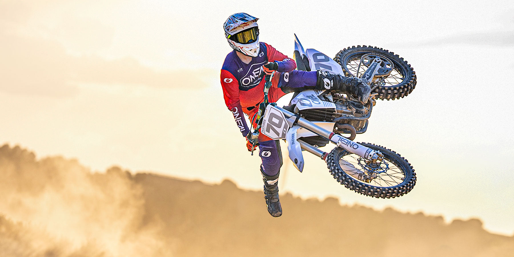 Ein Motocross-Fahrer in roter und blauer Kleidung vollführt einen Trick in der Luft auf einem Dirt Bike vor dem Hintergrund einer staubigen Landschaft und eines blassen Himmels.