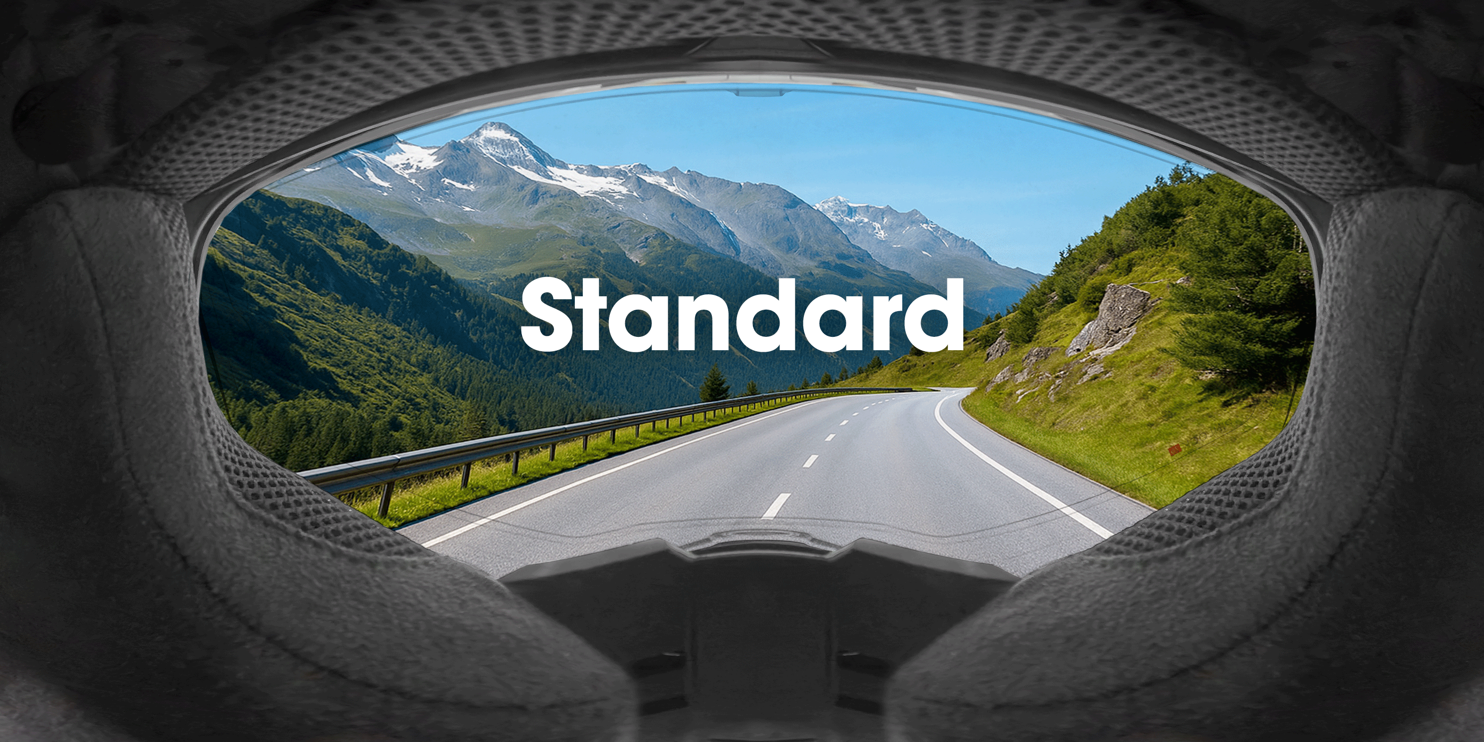 Blick auf eine kurvenreiche Bergstraße und grüne Hügel durch das Visier eines Motorradhelms, wobei das Wort "Standard" über das Bild gelegt ist.