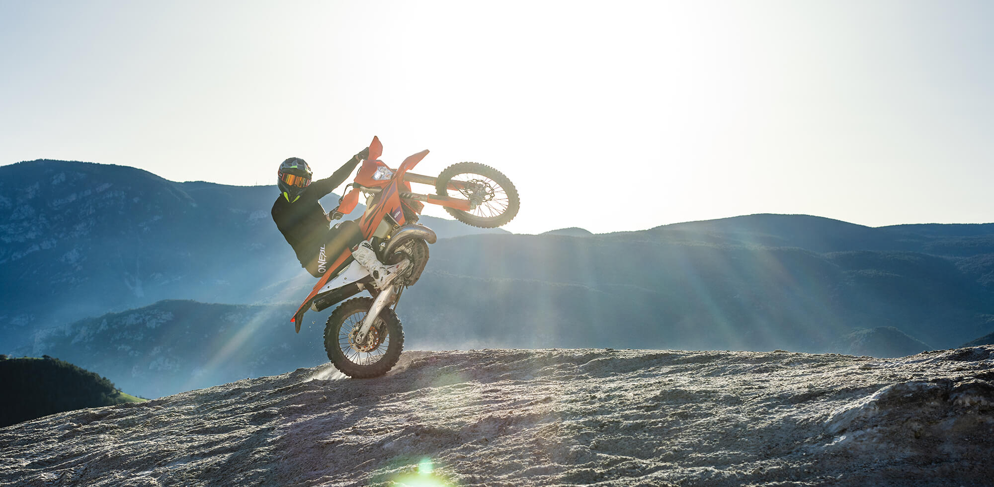 Eine Person fährt mit einem Dirtbike einen felsigen Hügel hinauf und macht dabei vor der Kulisse eines bergigen Geländes und eines hellen, sonnenbeschienenen Himmels einen Wheelie. Der Fahrer trägt einen Helm und Schutzkleidung, und das Sonnenlicht erzeugt einen dramatischen Linsenreflexionseffekt.